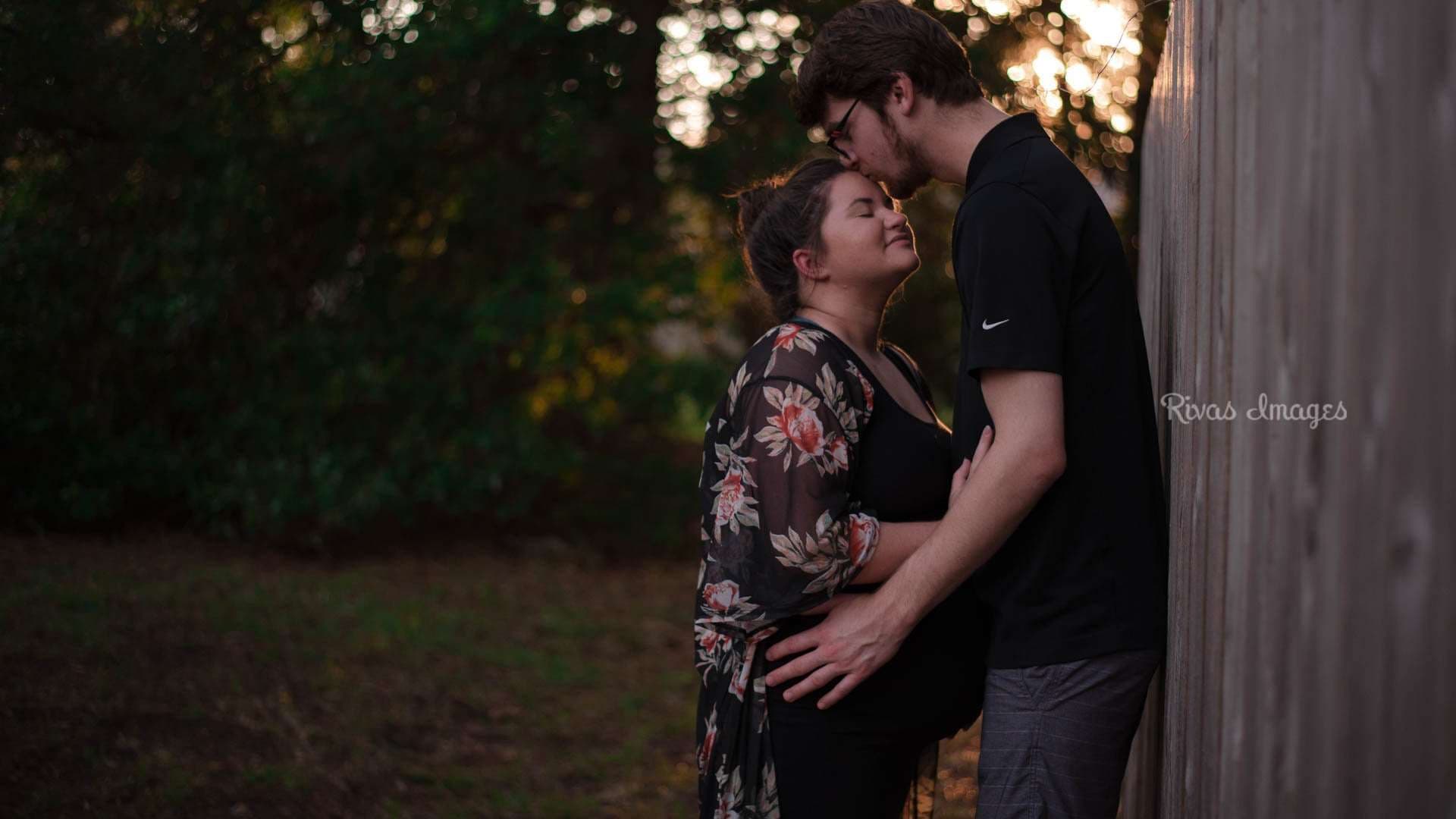 Tall man kissing his partner's forehead as they embrace beside a wooden fence at sunset, her hand on her belly
