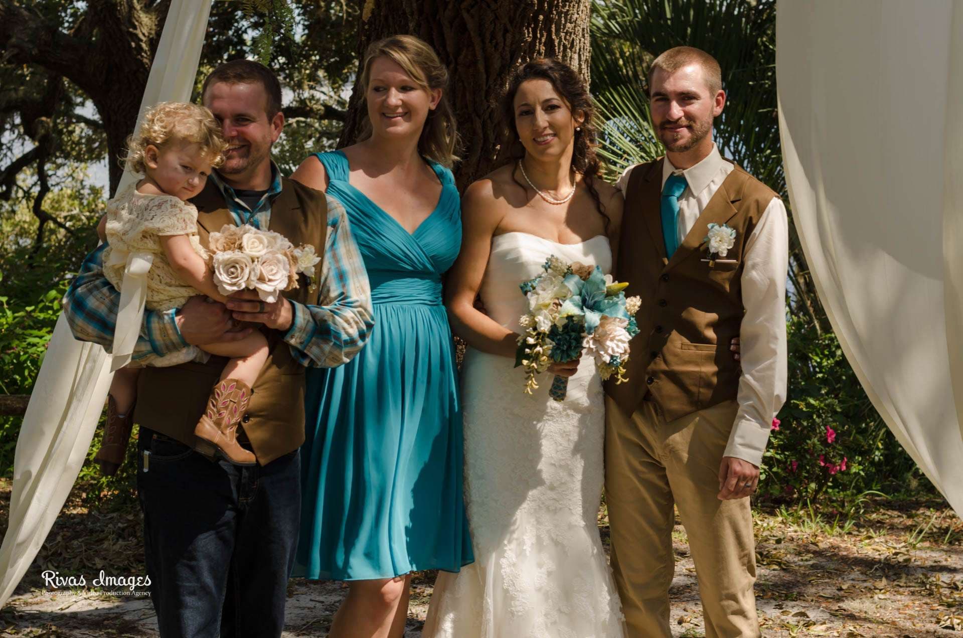 Bride in a strapless white gown holding a teal bouquet, posing with a groom, a bridesmaid in teal and a man holding a toddler