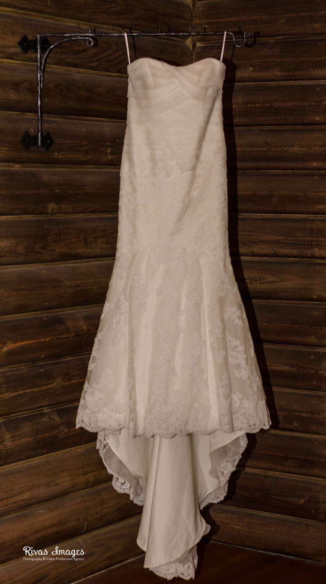 Strapless white lace mermaid wedding gown hanging from an iron hook against a dark rustic wooden wall