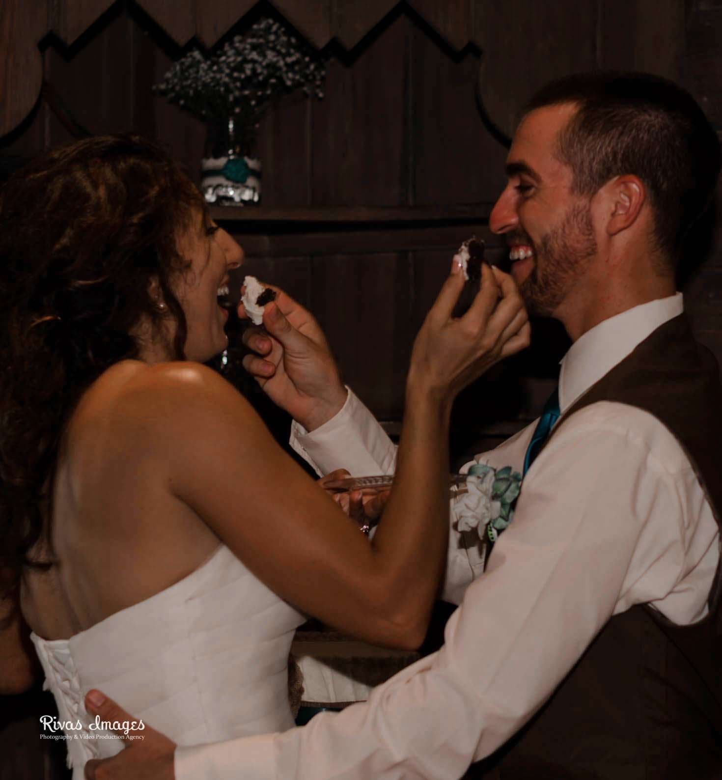 Bride and groom laughing as they feed each other wedding cake against a dark wooden wall