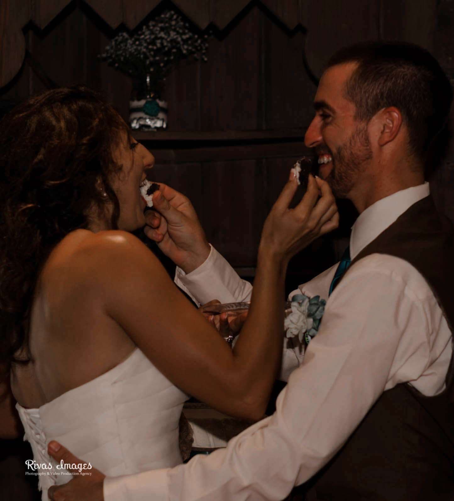 Couple laughing as they feed each other wedding cake at a rustic reception, bride in white strapless gown, groom in vest
