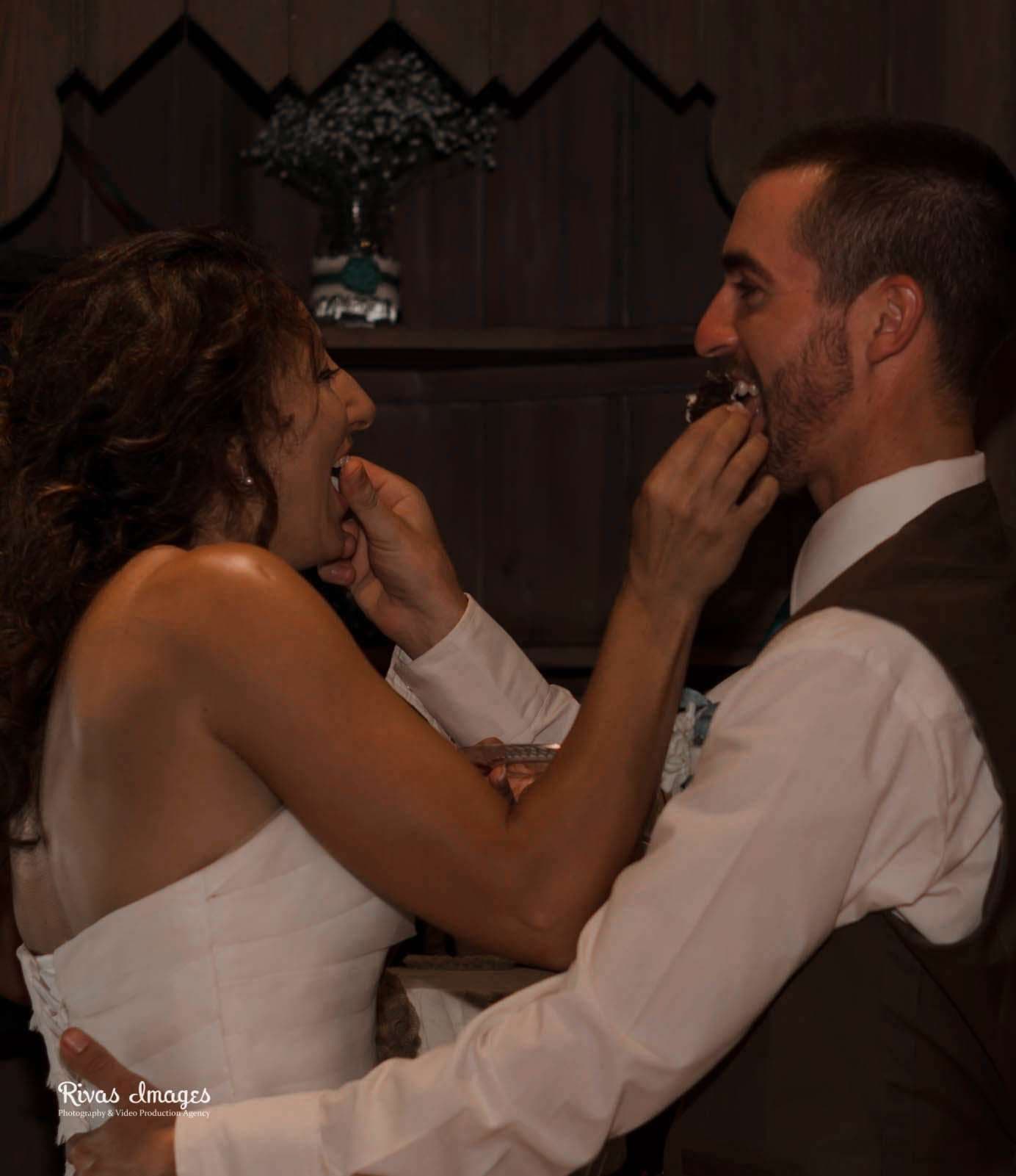Bride and groom laughing as they feed each other wedding cake in front of a dark wood wall