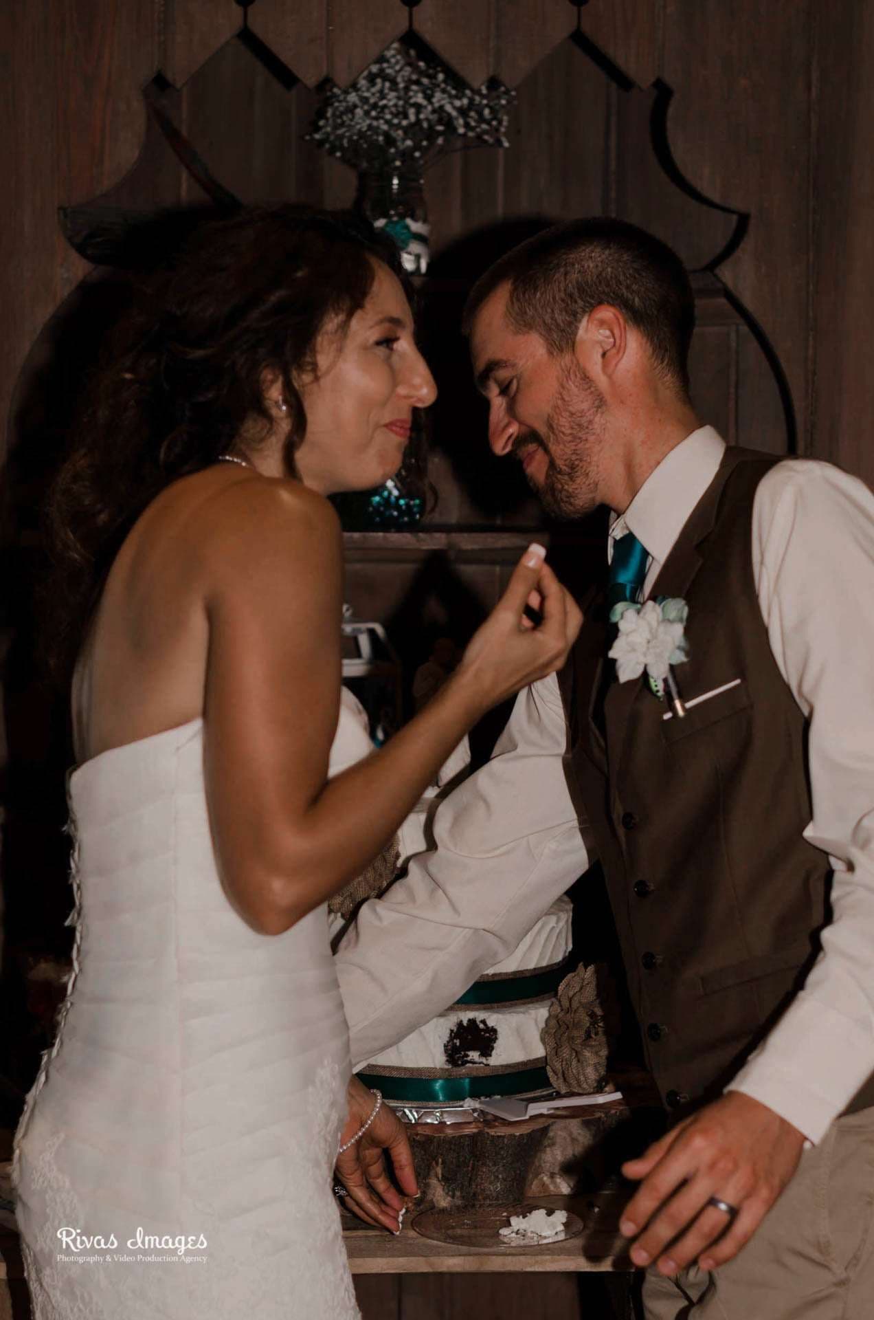 Bride in a strapless white gown feeds a bite of cake to a smiling groom in a brown vest and teal tie