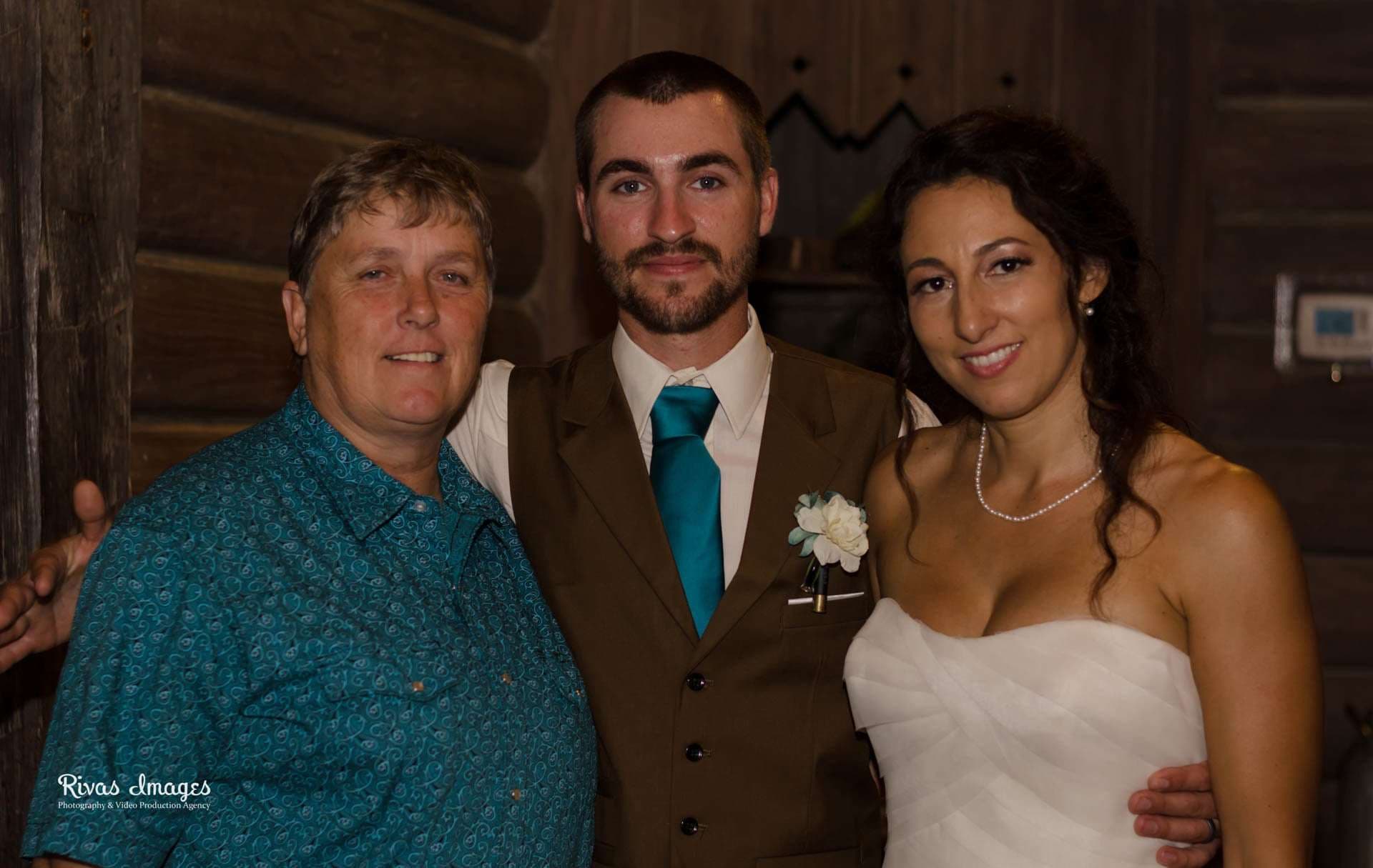 Bride in white gown and groom in brown vest with teal tie posing with a smiling guest in a teal shirt inside a log cabin