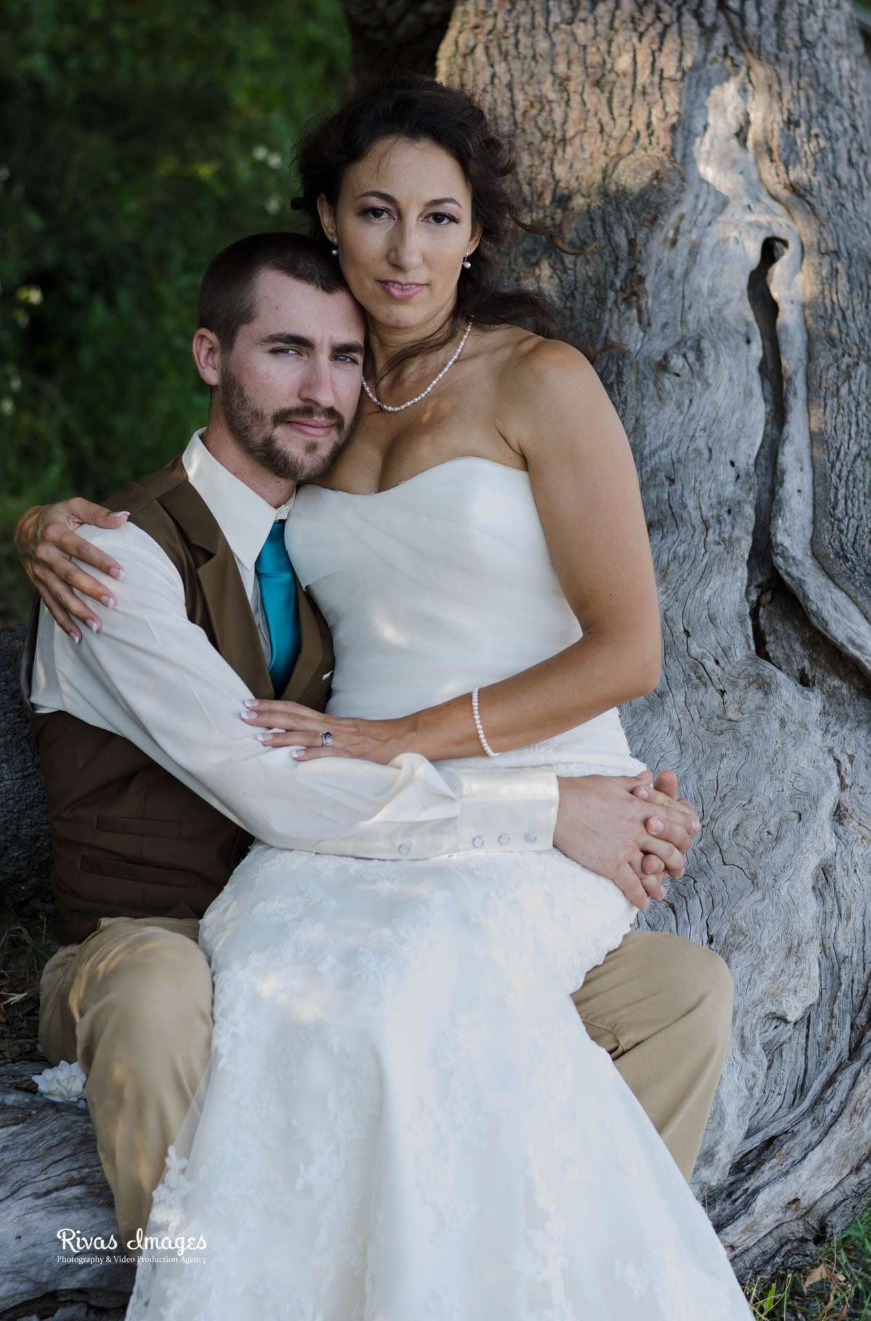 Groom in brown vest and teal tie embracing his bride in a strapless white gown beside a weathered tree