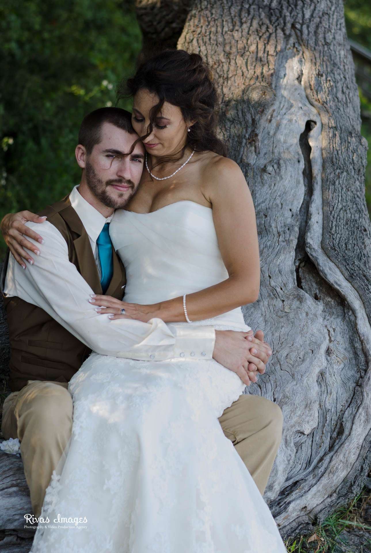 Bride in a strapless white gown sits on the groom's lap against a weathered tree trunk at Camp Helen State Park