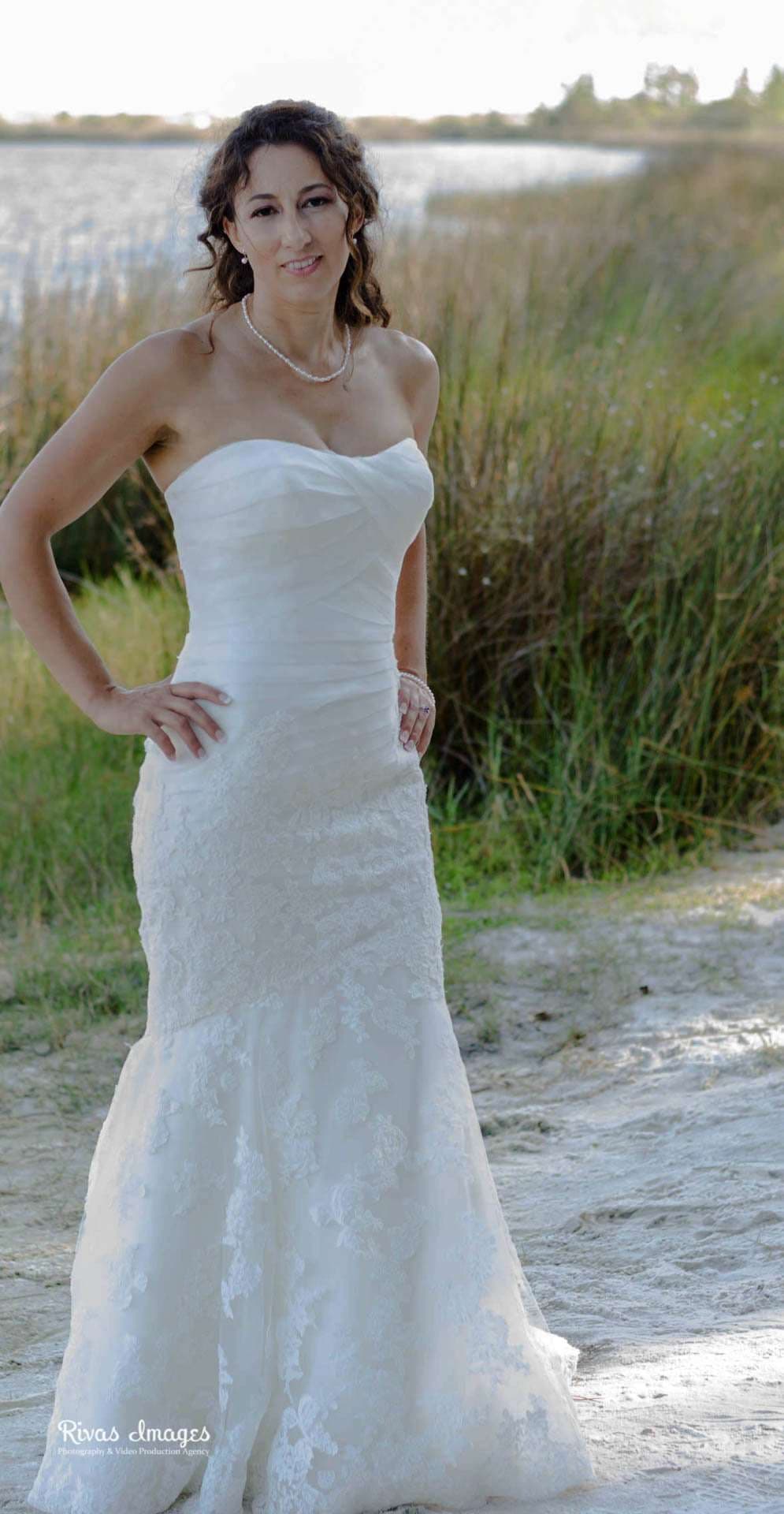 Bride in a strapless white lace gown with hand on hip, standing by tall marsh grass and a lake at Camp Helen State Park