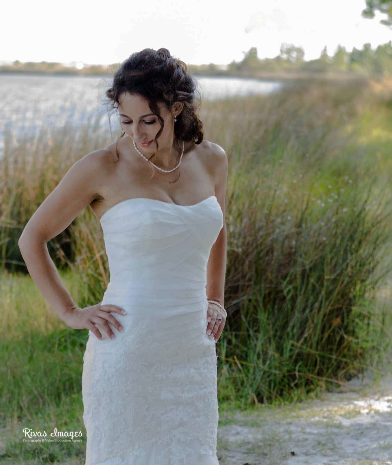 Bride in a strapless white lace gown and pearl necklace looks down with hand on hip beside tall lakeside grass