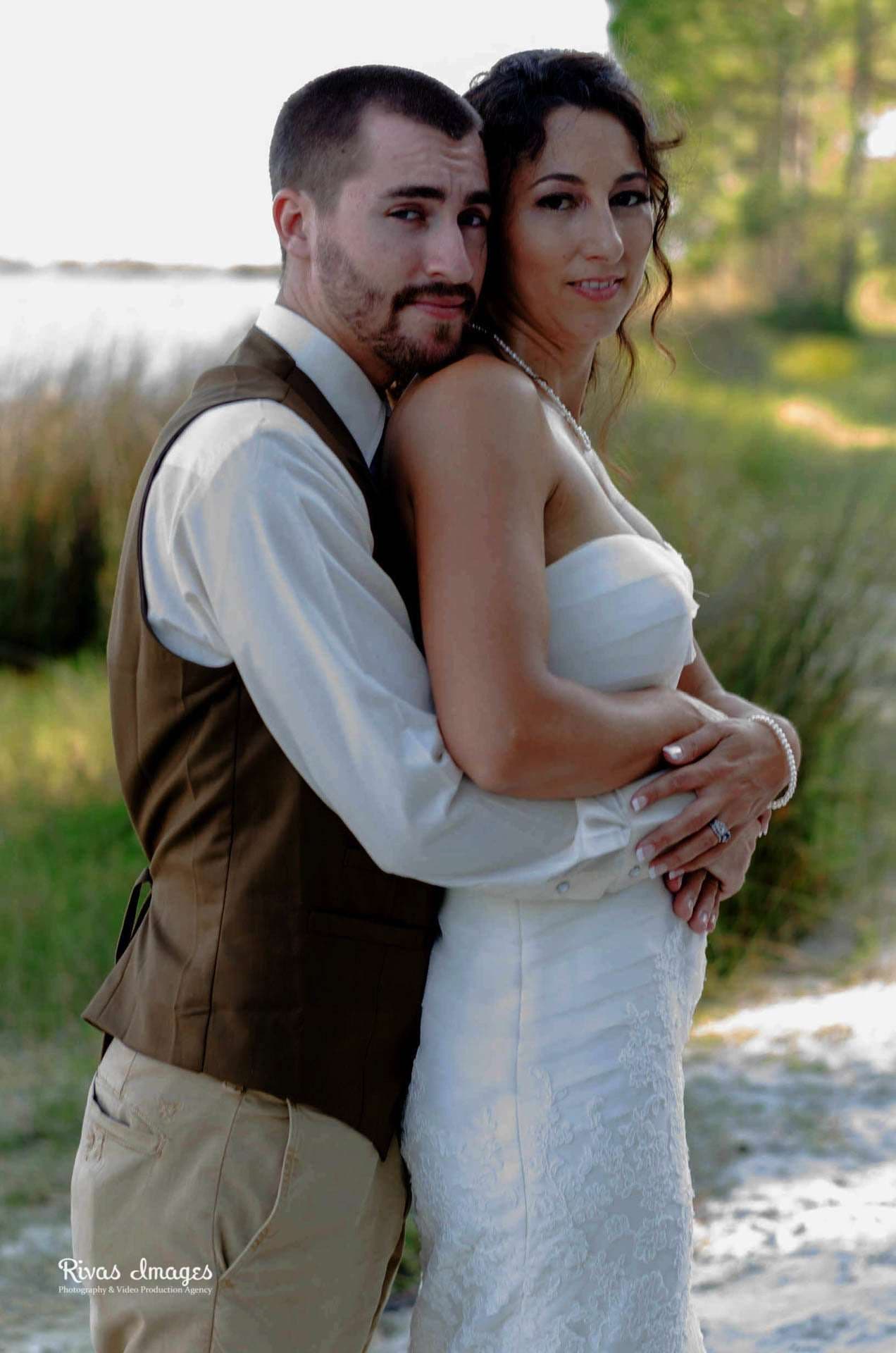 Groom in brown vest embracing his bride from behind at Camp Helen State Park, lake and tall grass behind them