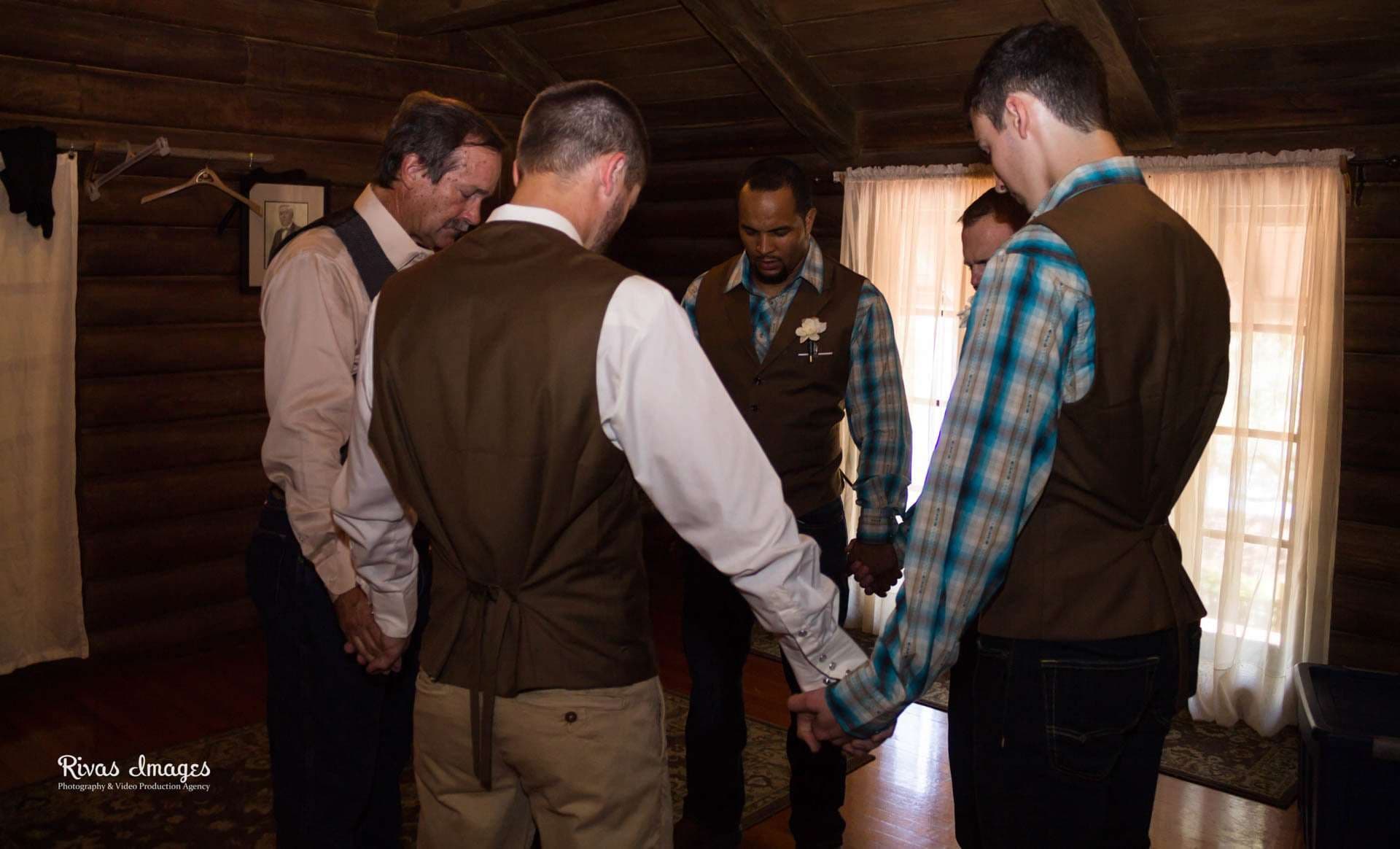 Four men in brown vests and plaid shirts stand in a circle holding hands with heads bowed inside a log cabin