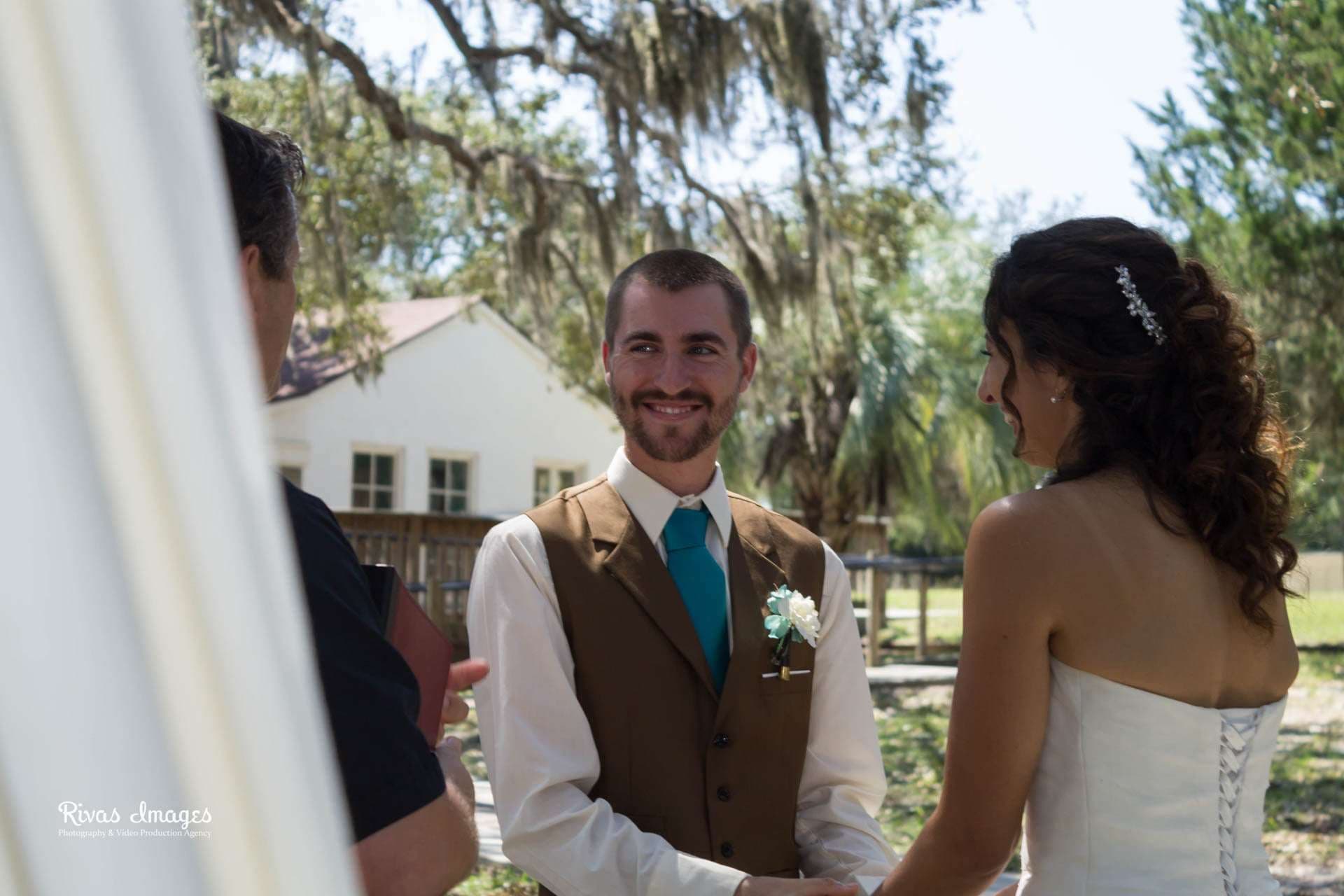 Smiling groom in brown vest and teal tie holds hands with his bride during an outdoor ceremony at Camp Helen State Park