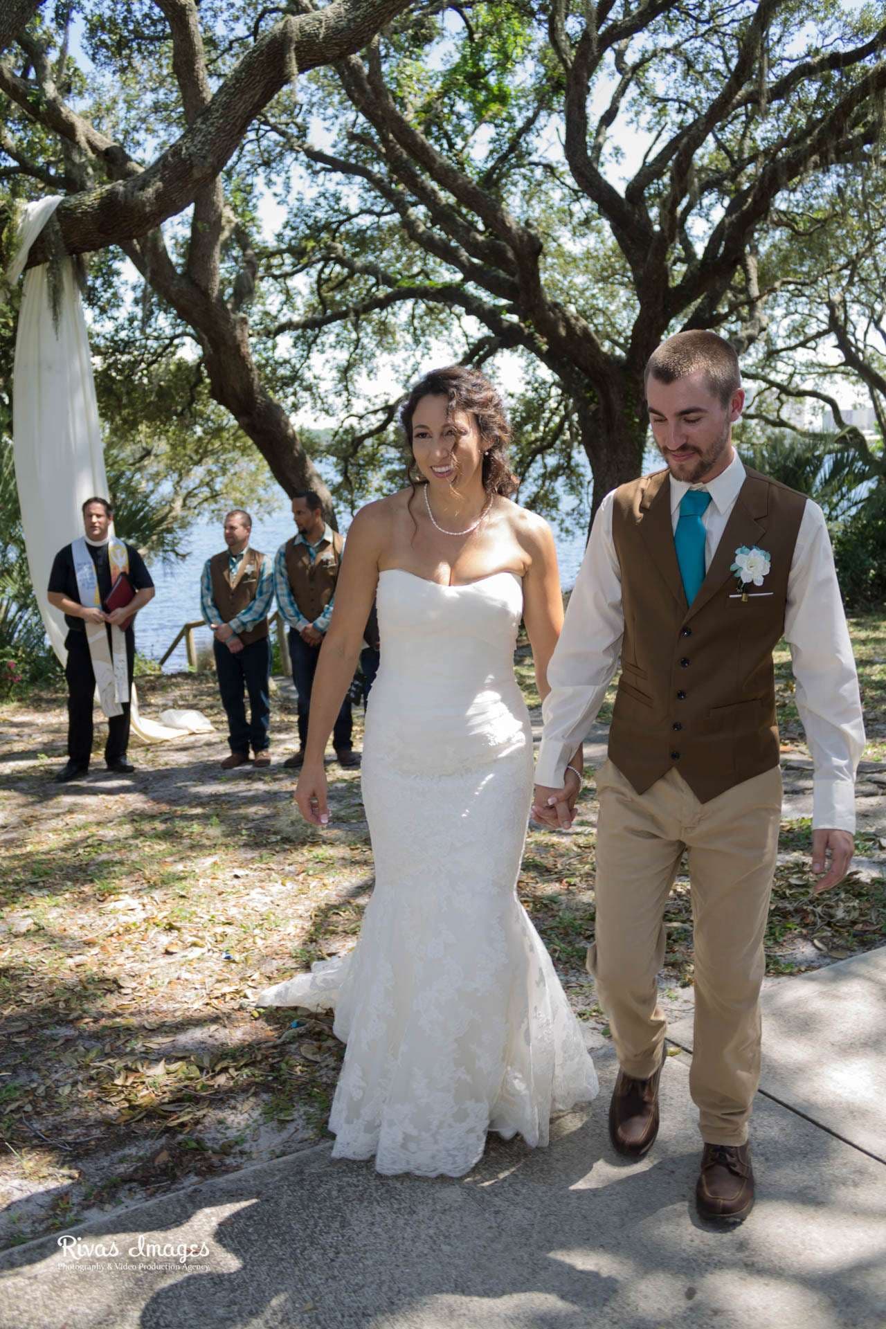 Smiling bride in a white lace gown holding hands with groom in brown vest under oak trees at Camp Helen State Park