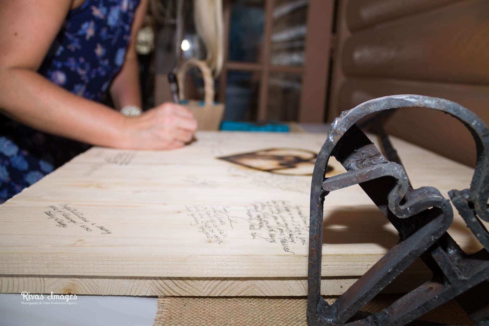 Guest signing a wooden board guest book with a pen beside a heart-shaped branding iron at a rustic wedding