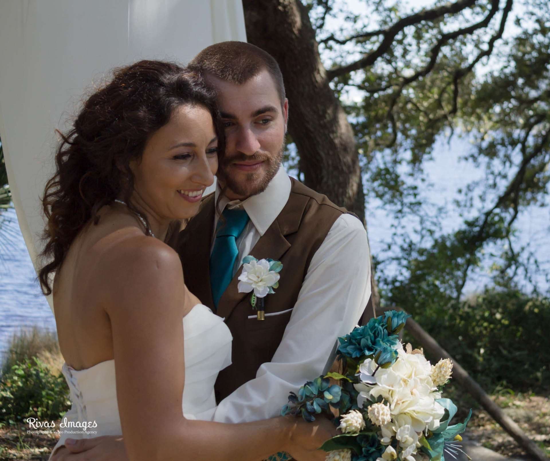 Smiling bride in white gown embraced by groom in brown vest and teal tie, holding teal and white bouquet by the water