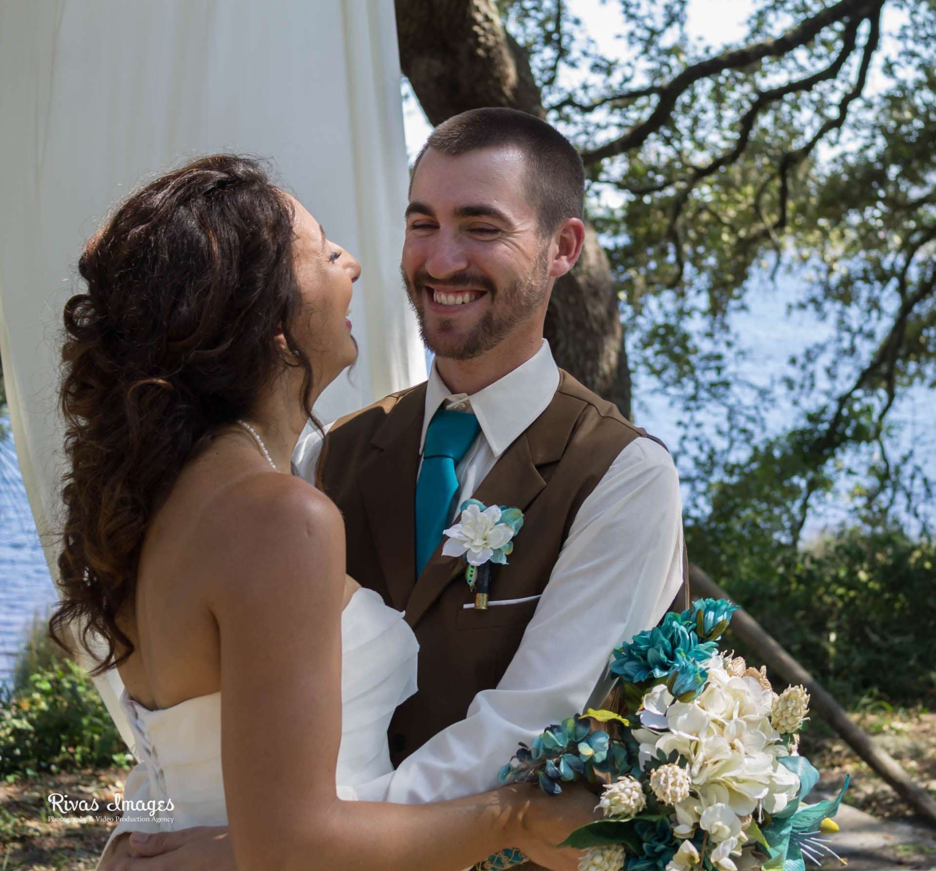 Laughing couple embracing under an oak tree by the water, groom in brown vest and teal tie, bride holding teal bouquet