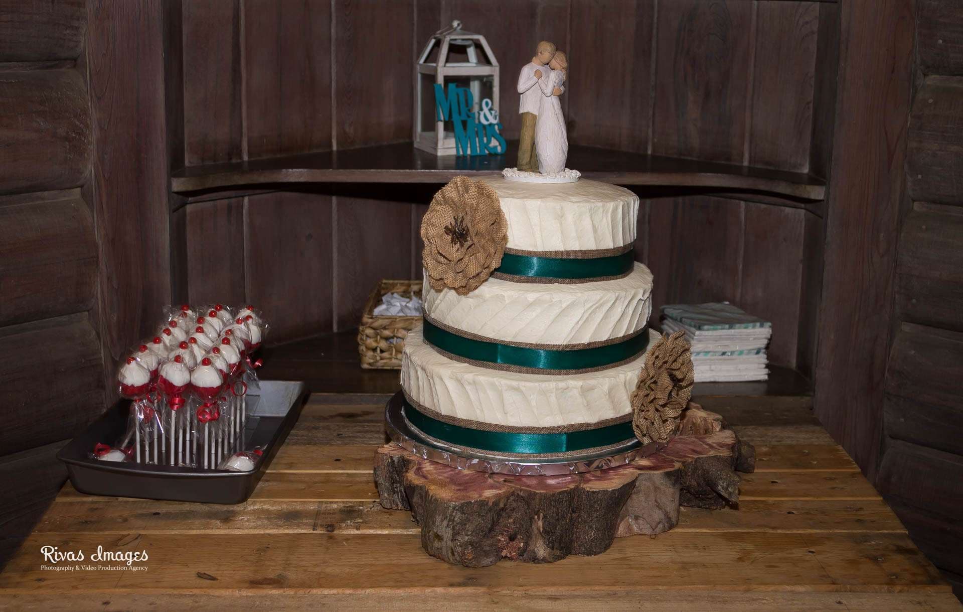 Three-tier white wedding cake with teal ribbon on a rustic log slice stand, beside red and white cake pops