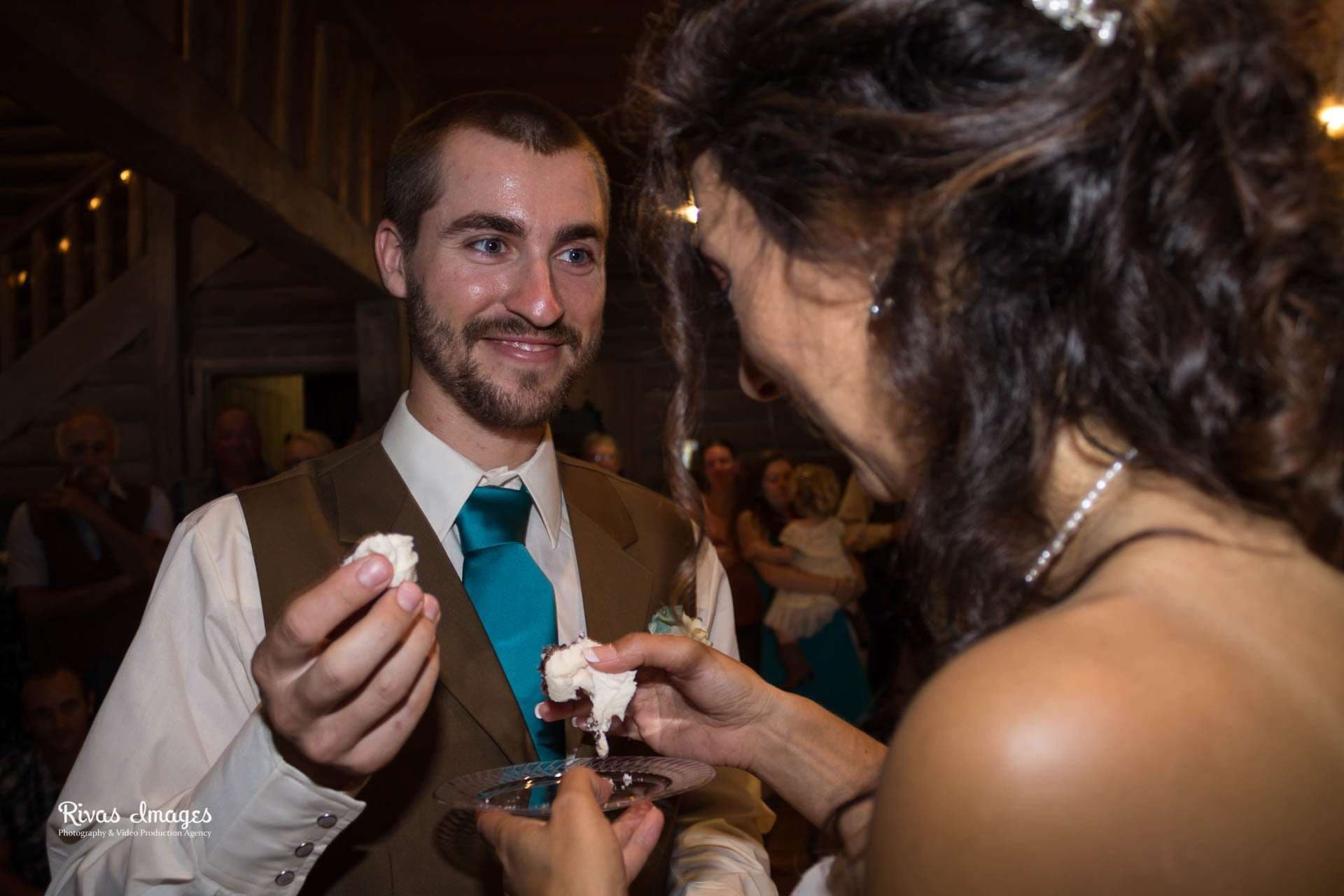 Smiling groom in a brown vest and teal tie holds a piece of cake while sharing cake with his bride
