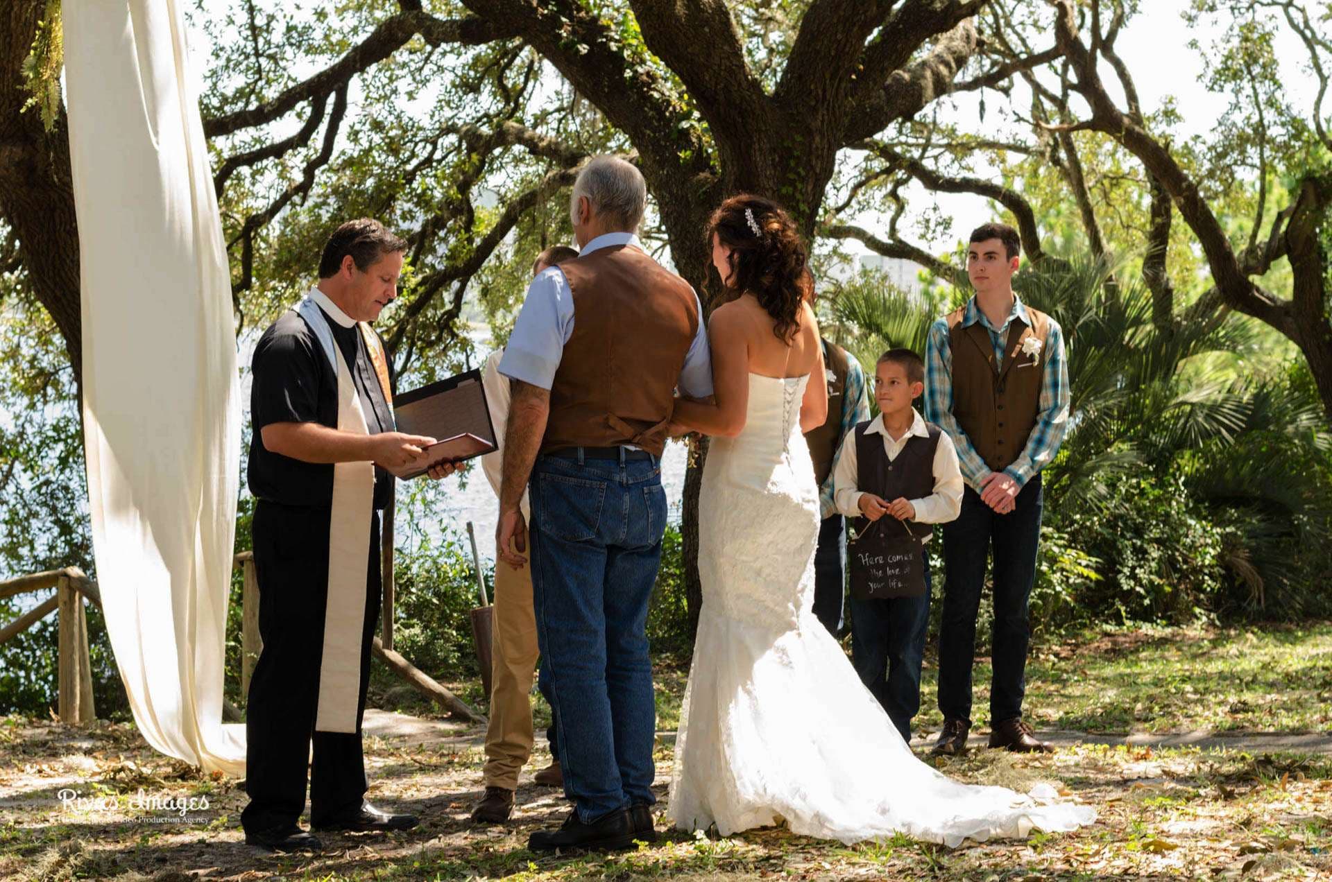 Officiant reading to a bride in a white gown and a man in a leather vest under a large oak at Camp Helen State Park