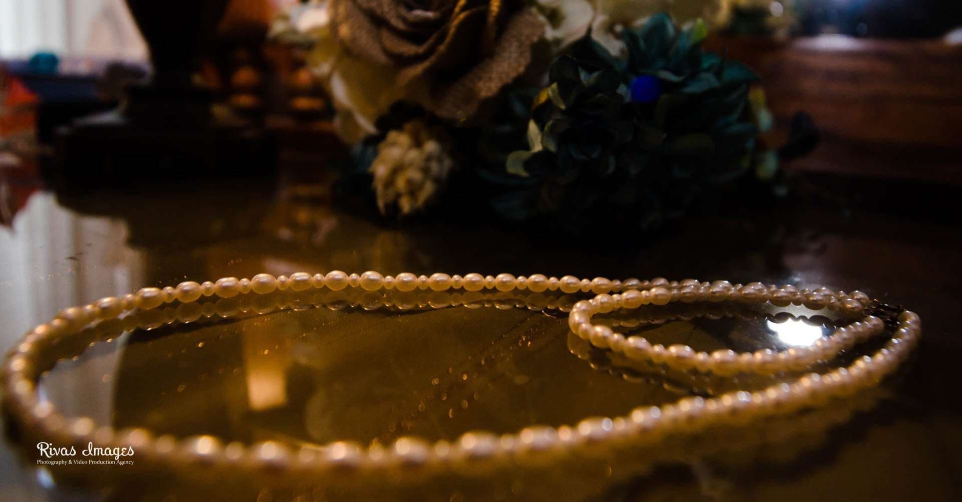 Strand of pearls resting on a glossy wooden table in front of a blurred burlap and teal flower bouquet