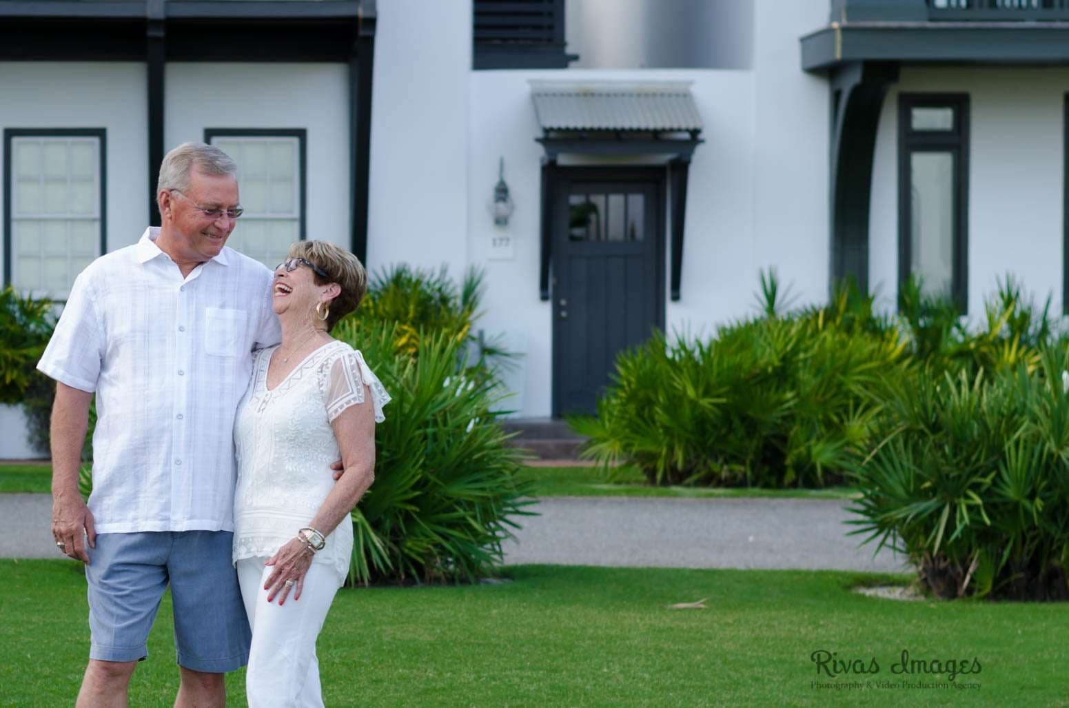 Older couple laughing and embracing on a lawn in front of a white house with green shrubs in Rosemary Beach