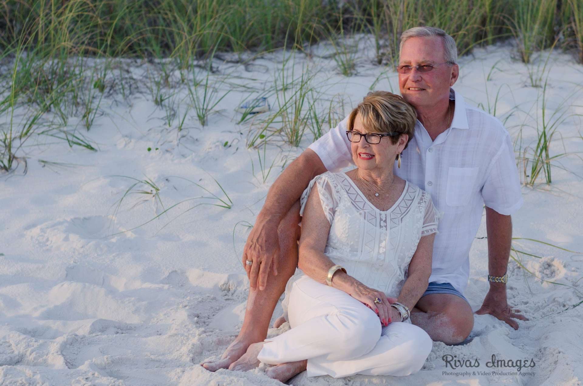 Older couple in white sitting close together on a sandy beach with dune grass behind them in Rosemary Beach