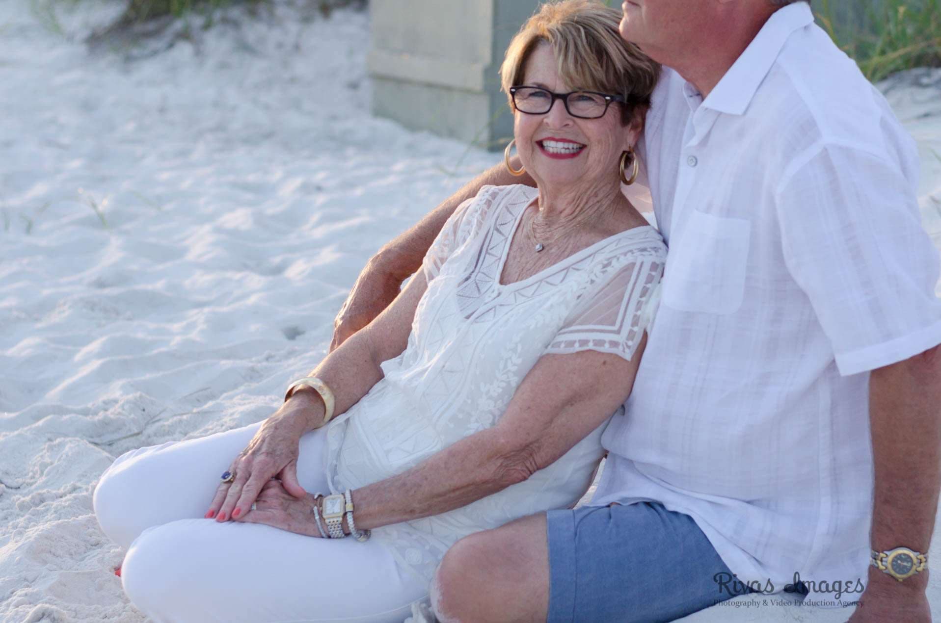 Smiling older woman in white lace top and glasses sitting in the sand, leaning against a man in a white shirt