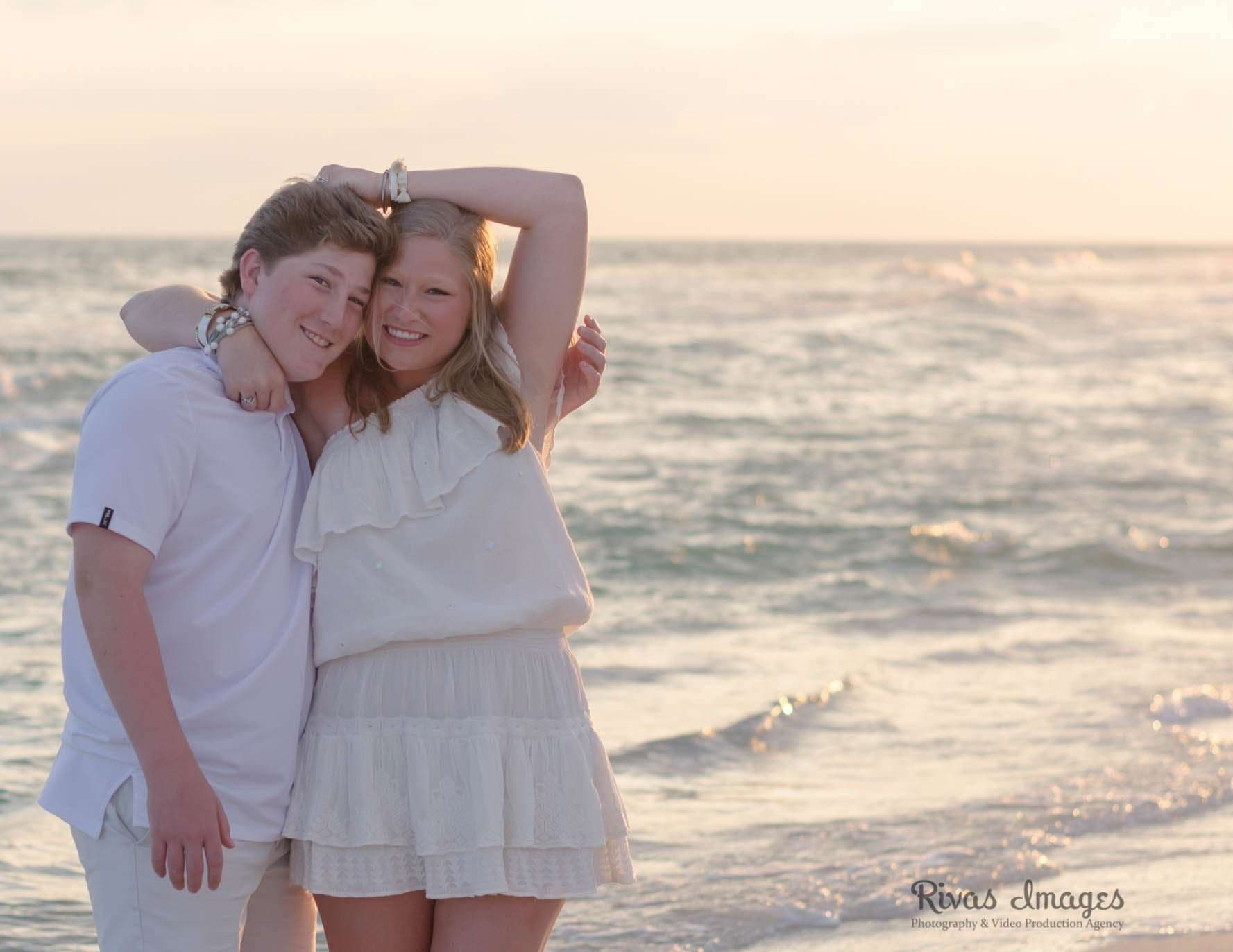Two smiling young people embracing on the beach at sunset, one in a white top and skirt, waves behind them