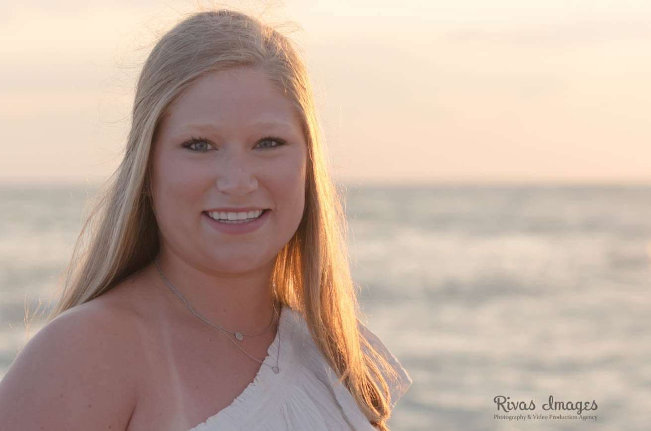 Smiling woman with long blonde hair in a white one-shoulder top, backlit by a soft sunset over the water