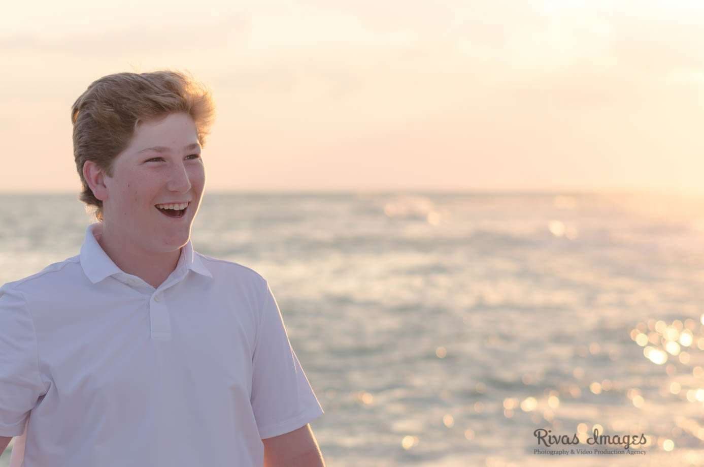 Smiling teenager in a white polo laughing beside the sparkling ocean at sunset in Rosemary Beach
