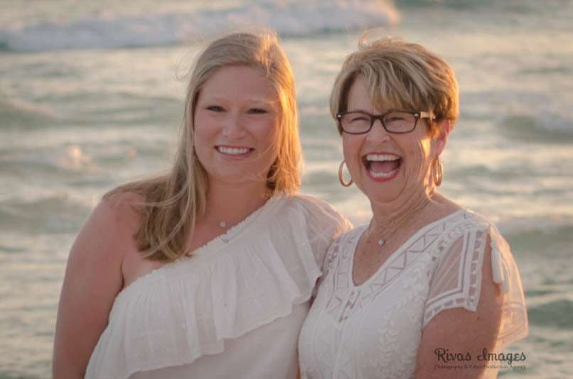 Two smiling women in white tops laughing together on the beach at sunset, with ocean waves behind them