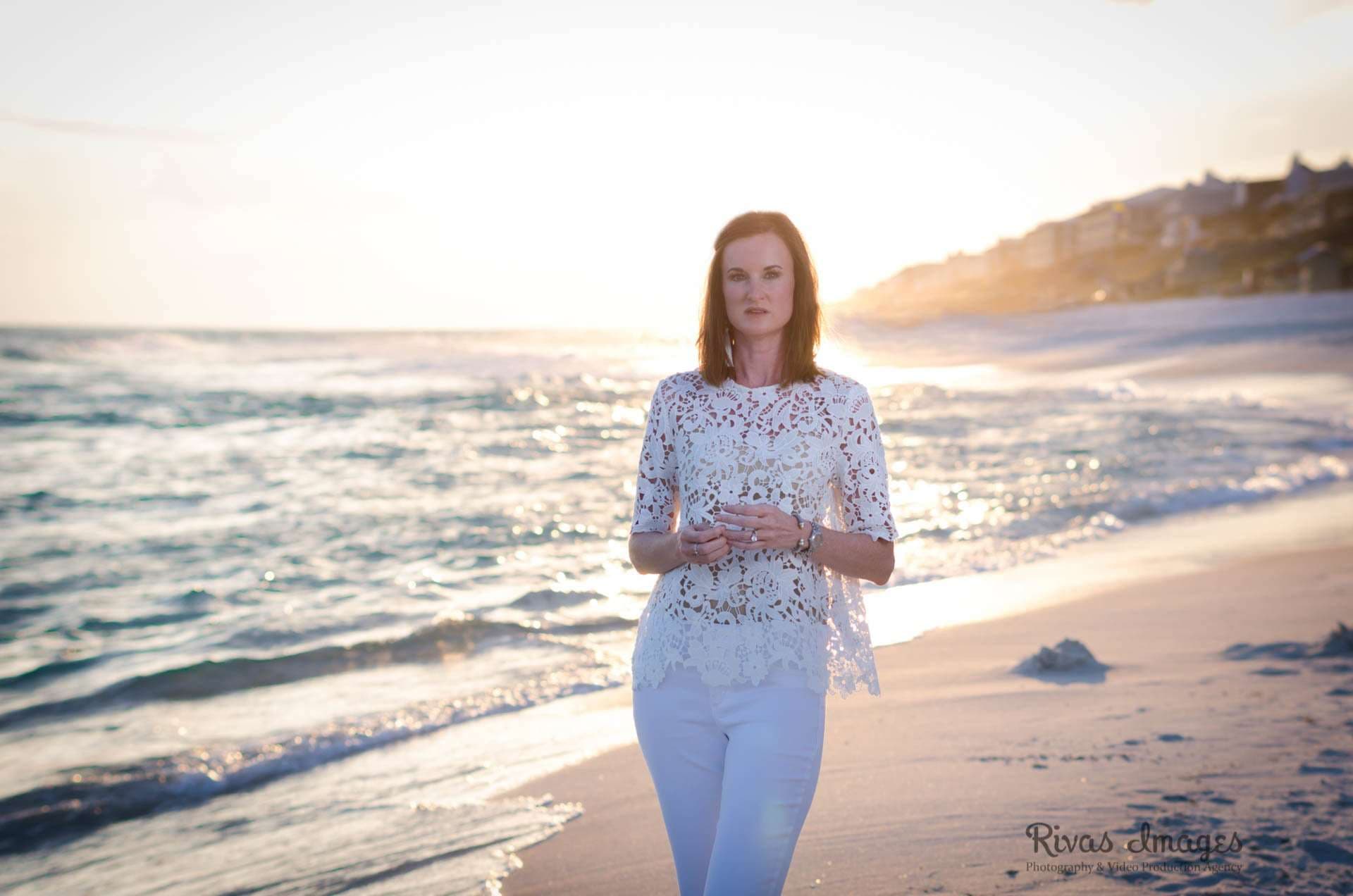 Woman in a white lace top and white pants standing on the shore at Rosemary Beach at sunset