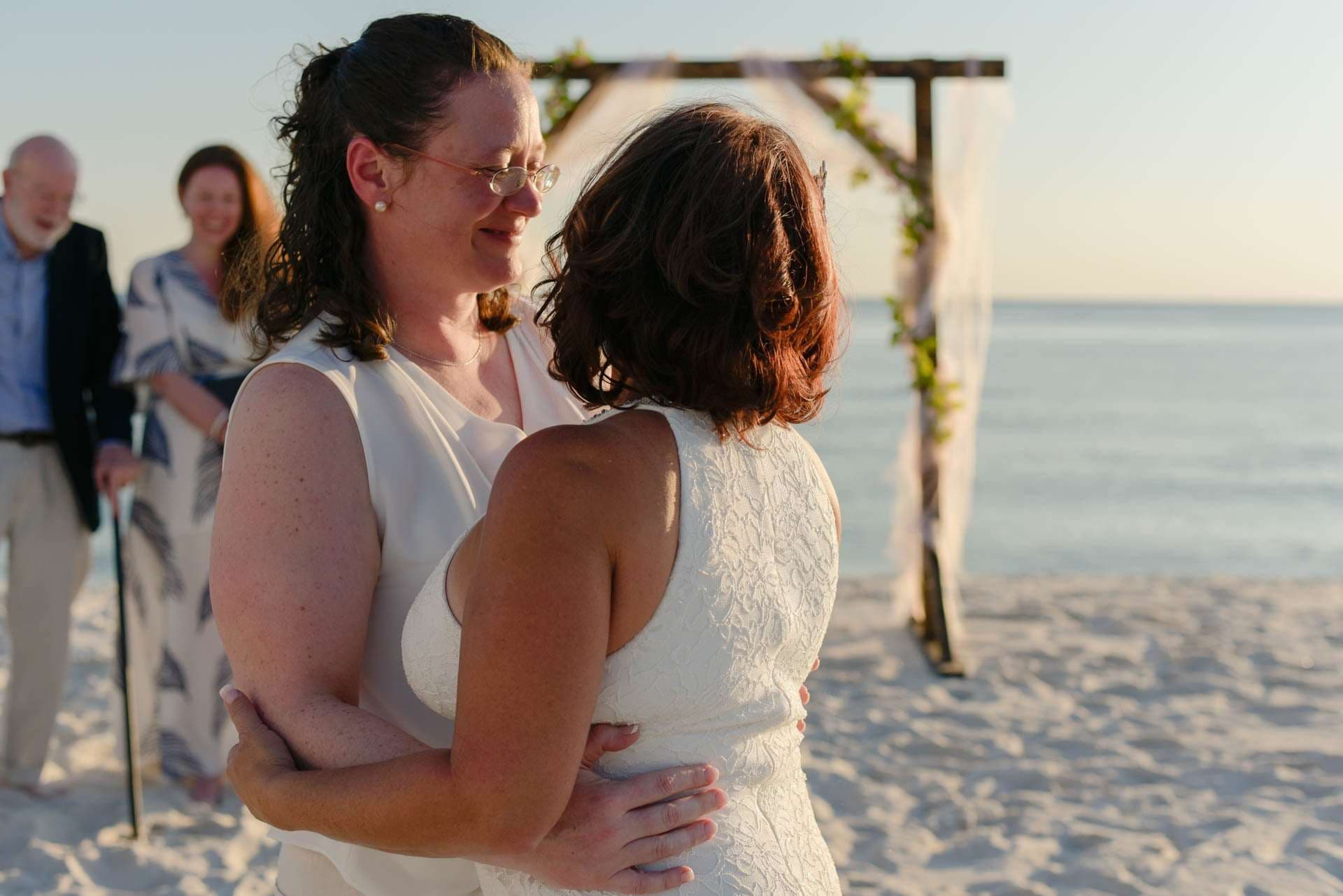 Two women in white embracing on the beach beside a floral wedding arch while a smiling guest looks on