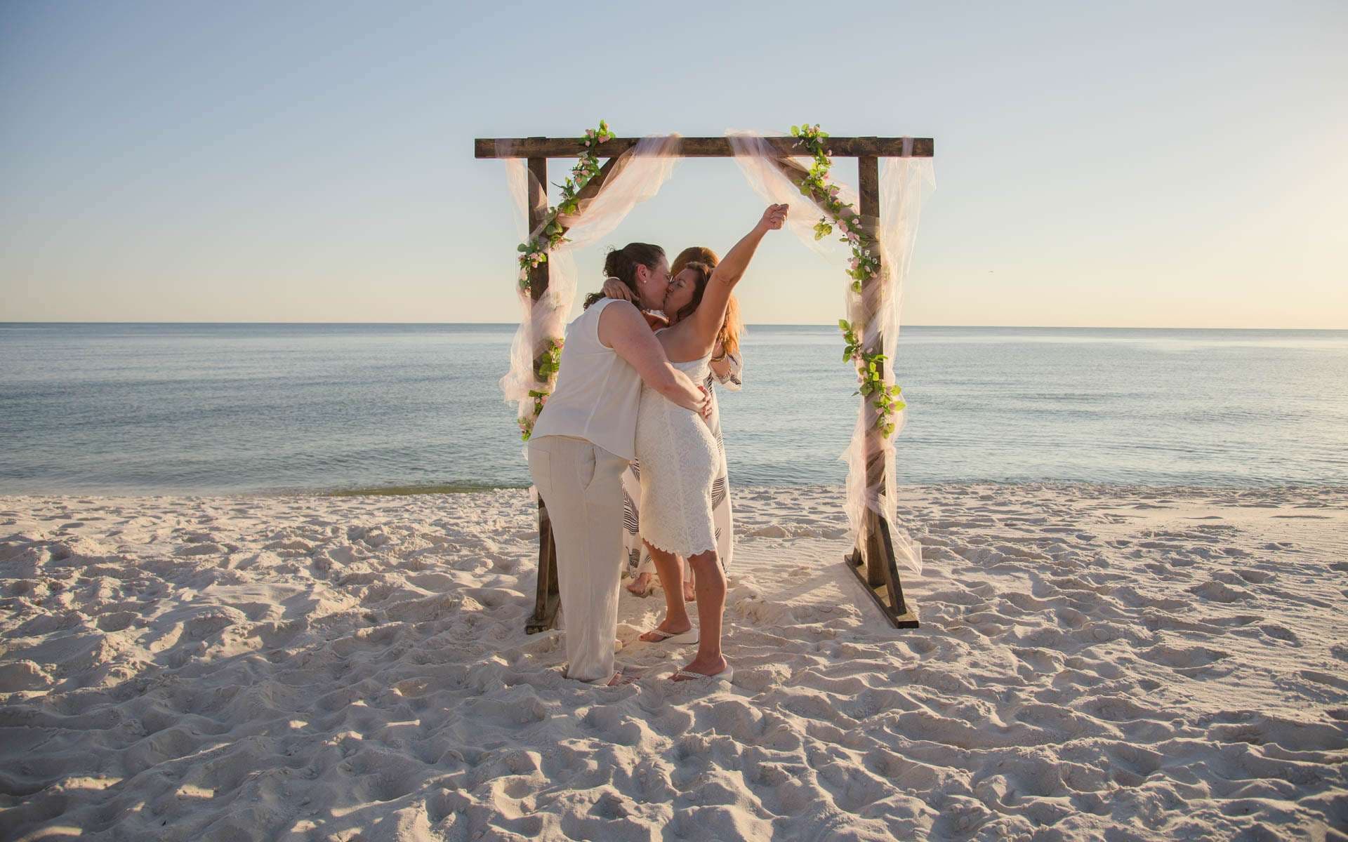 Couple kissing under a wooden wedding arch draped with greenery and tulle on a white sand beach at sunset