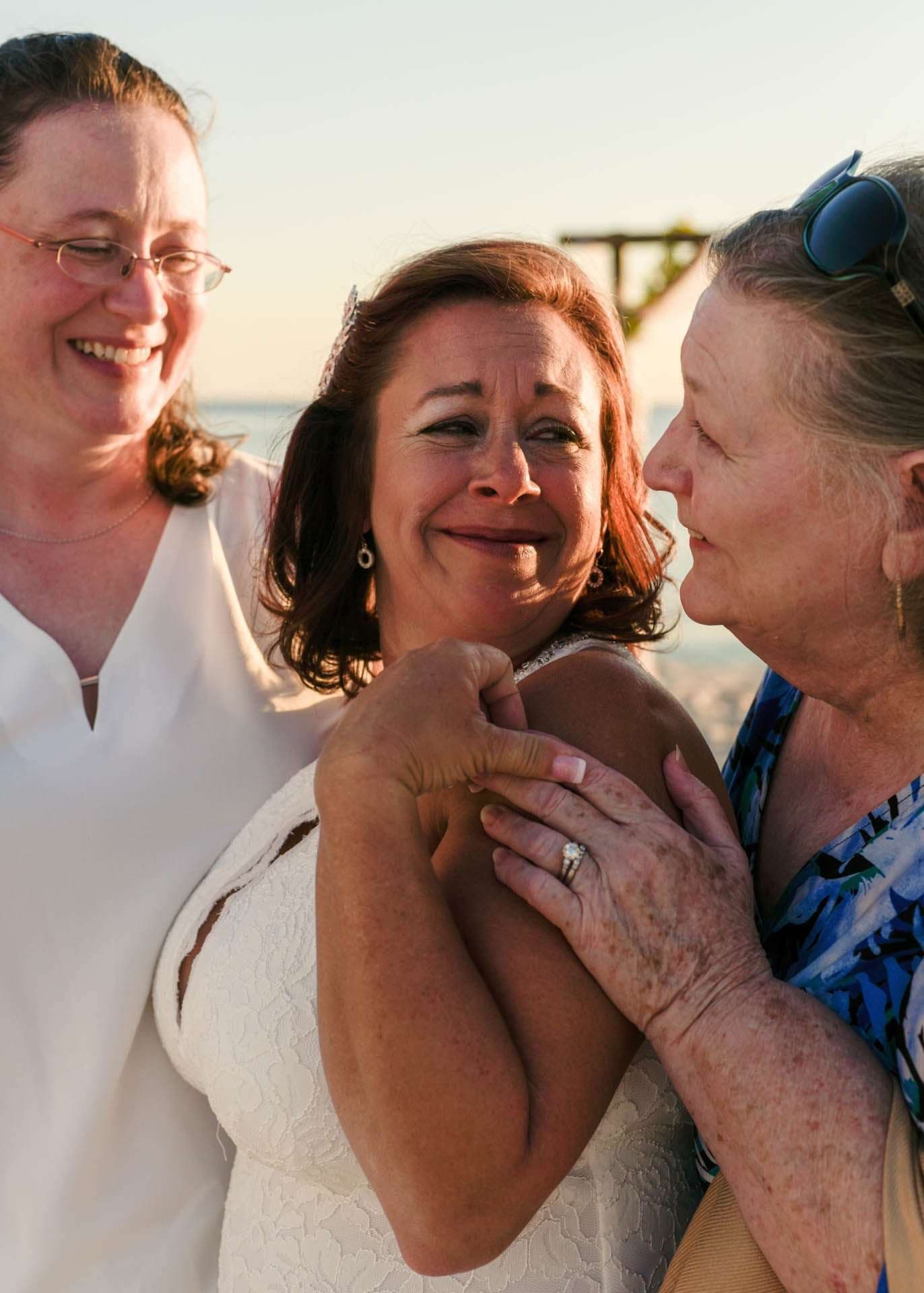 Smiling bride in white lace dress embraced by two women on the beach at sunset