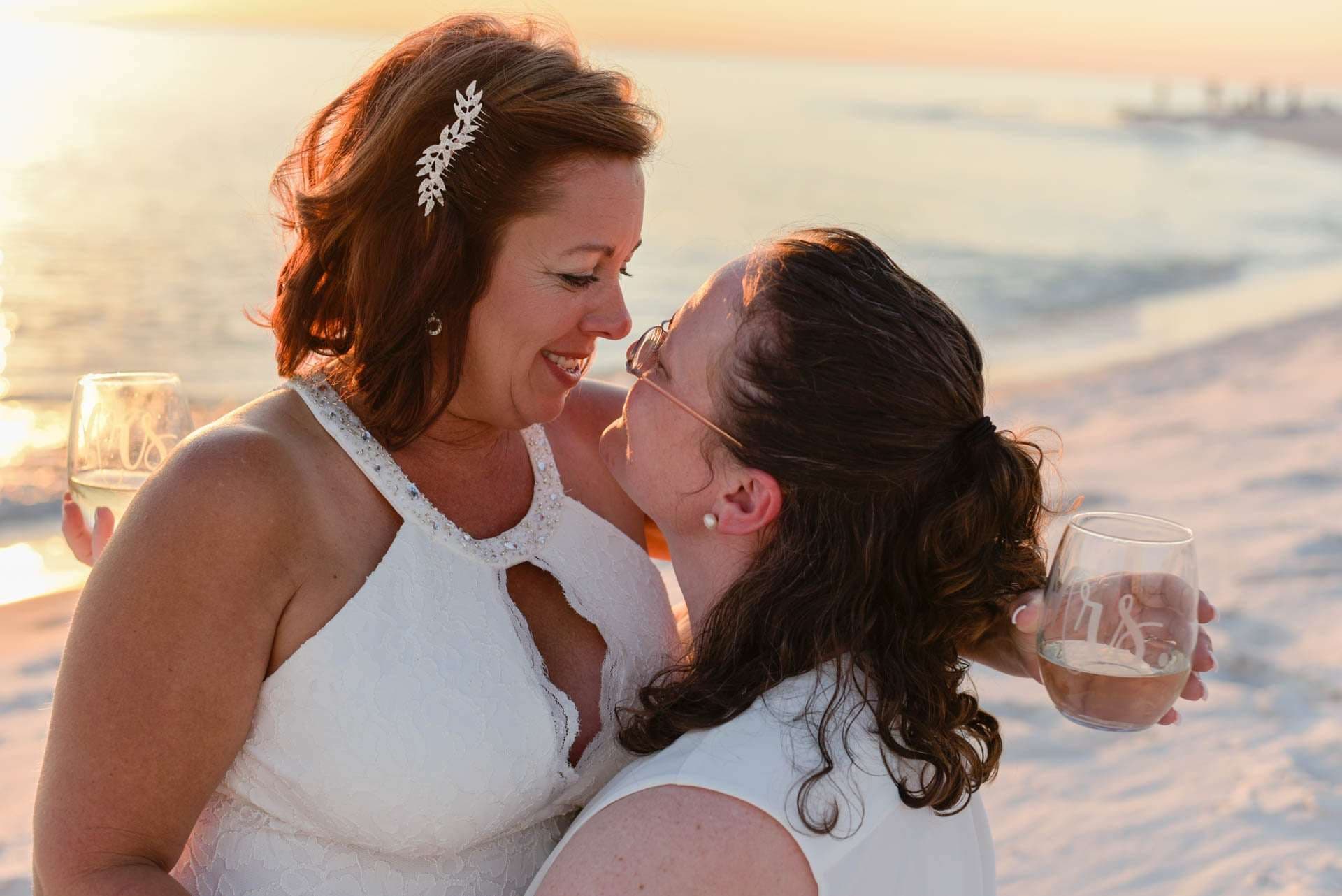Two brides in white nearly touching noses on the beach at sunset, holding wine glasses