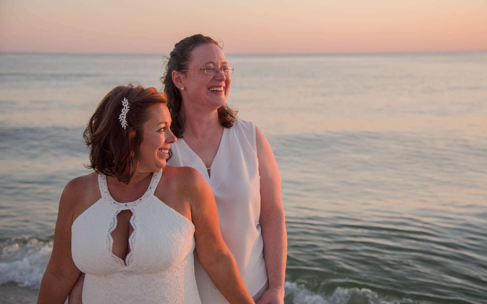 Two women in white outfits laughing and looking out over the water on the beach at sunset