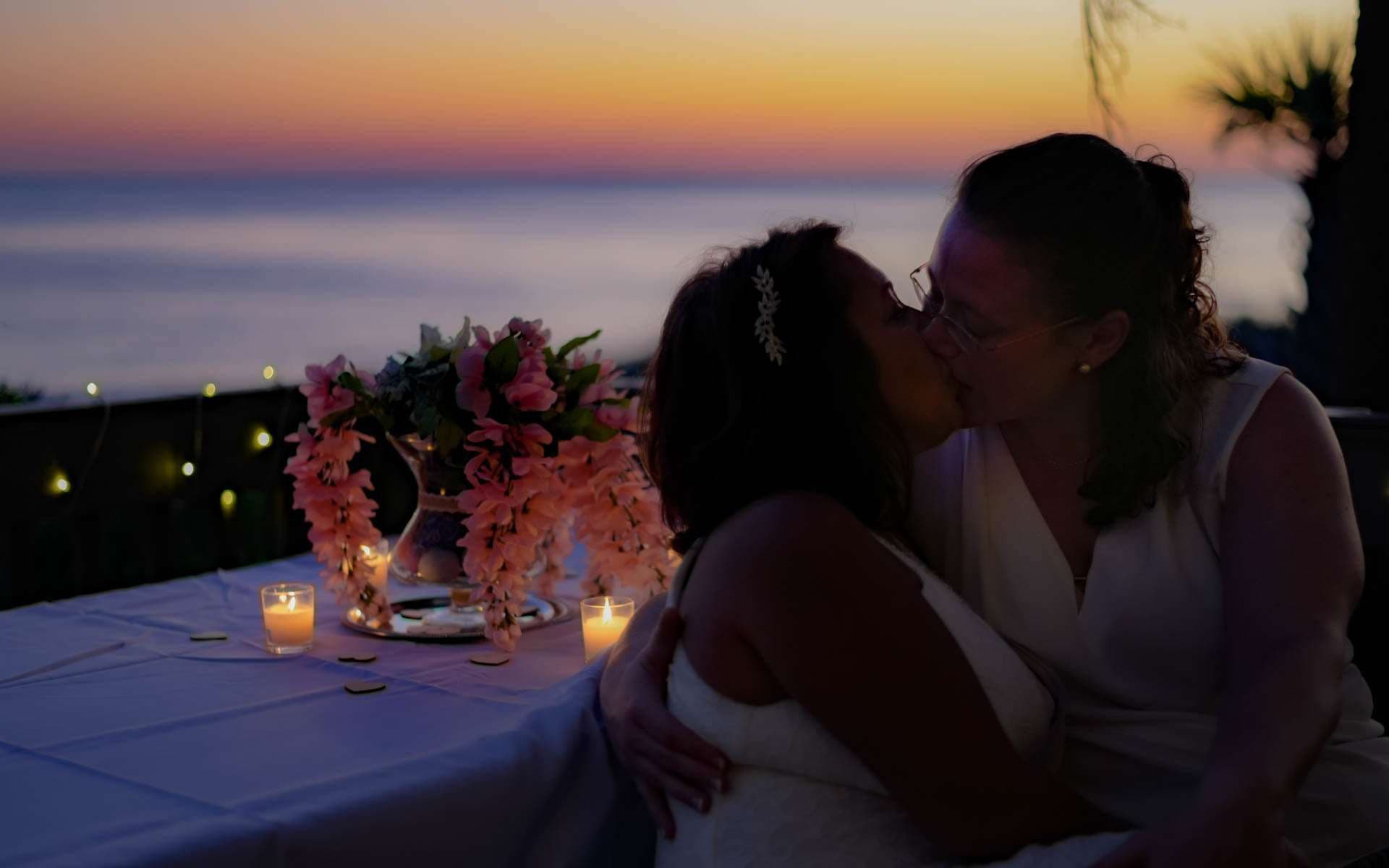 Two brides in white kissing on a terrace at sunset beside a candlelit table with pink floral centerpiece and ocean view