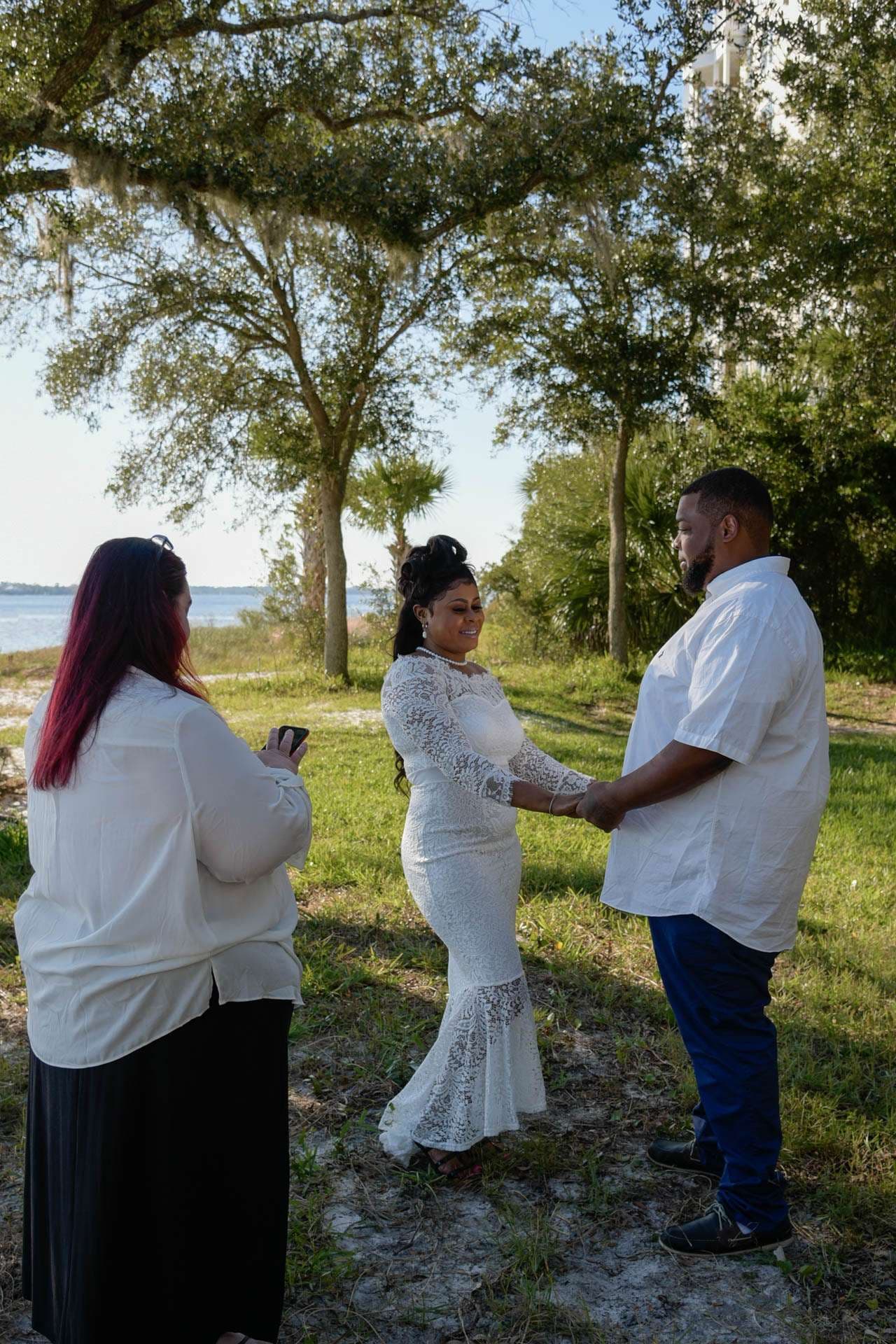 Couple in white holding hands during an outdoor elopement ceremony under oak trees by the water, officiant facing them