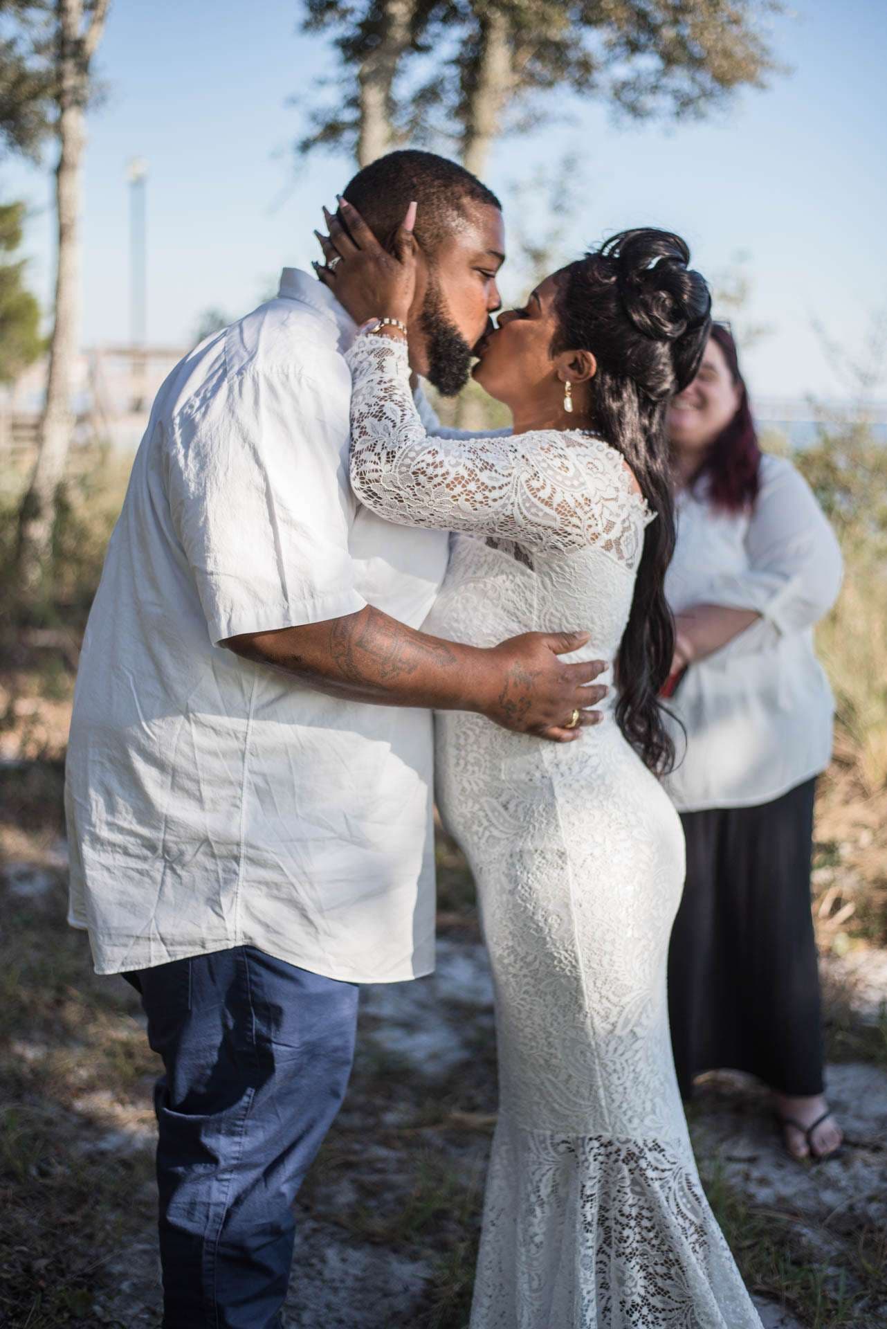 Couple kissing outdoors during their elopement ceremony, bride in a white lace gown, officiant smiling behind them