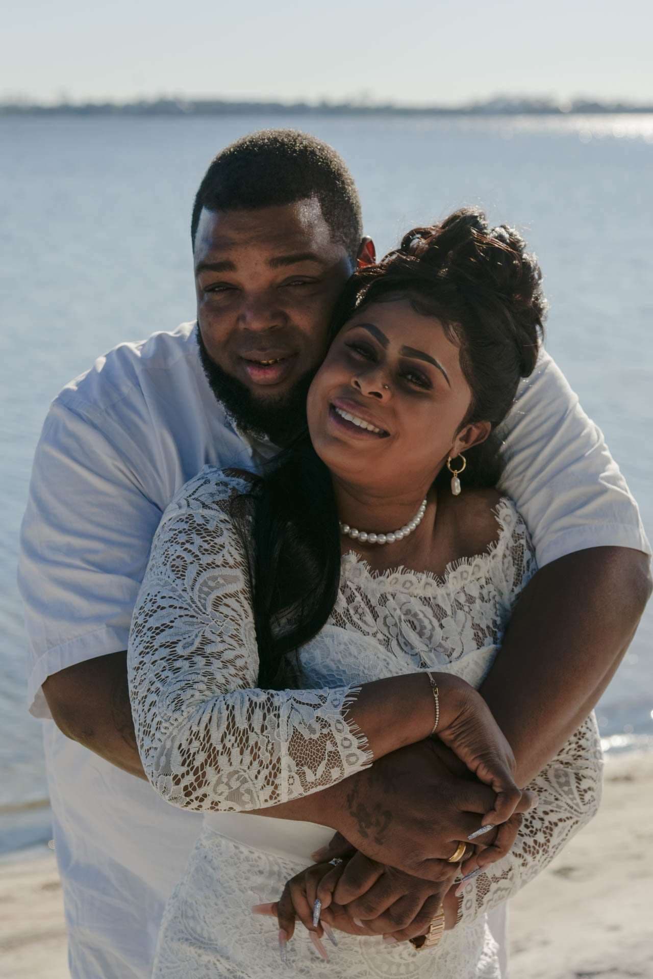 Smiling couple embracing by the water, man hugging woman in white lace dress and pearl necklace from behind