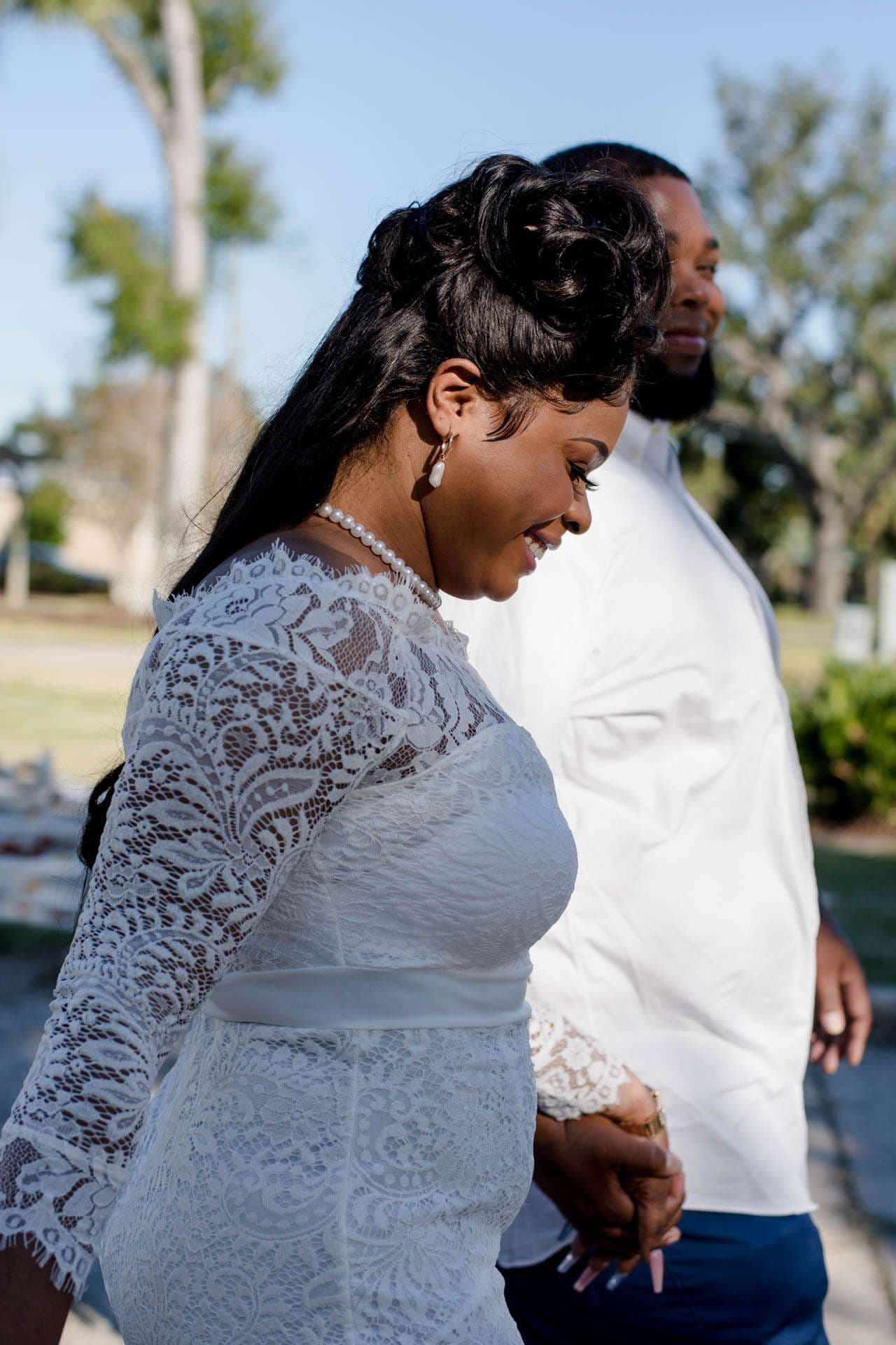 Smiling bride in a white lace dress walking hand in hand with her groom in a white shirt through a sunny park