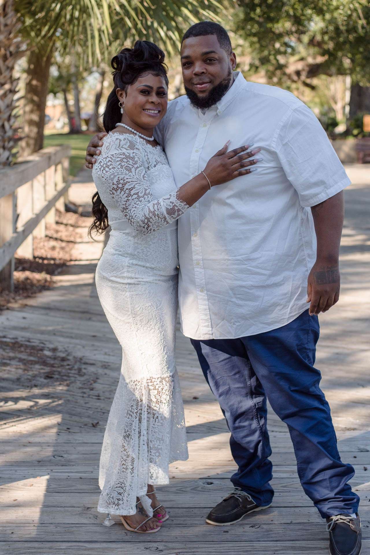 Smiling couple embracing on a wooden boardwalk, she in a white lace gown, he in a white shirt and blue pants
