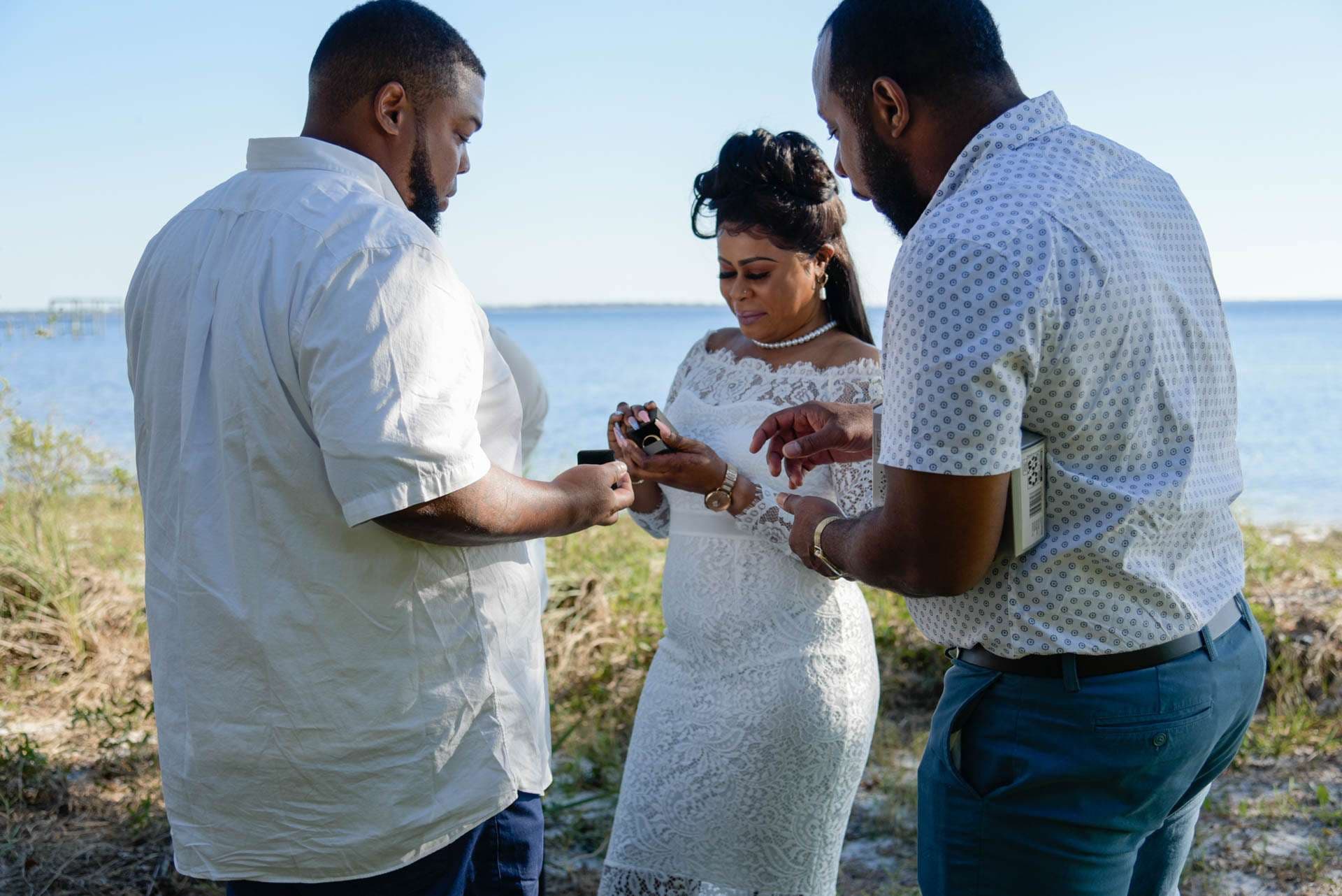 Bride in white lace dress looks at a ring while two men in button-up shirts stand beside her by the bay