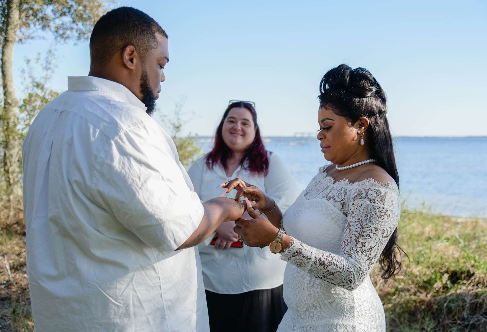 Couple exchanging rings during an outdoor ceremony at Oaks By The Bay, officiant smiling behind them, water in background