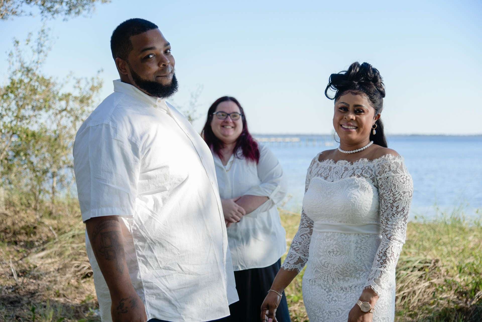 Couple in white outfits smiling by the bay, with a smiling officiant in glasses standing behind them