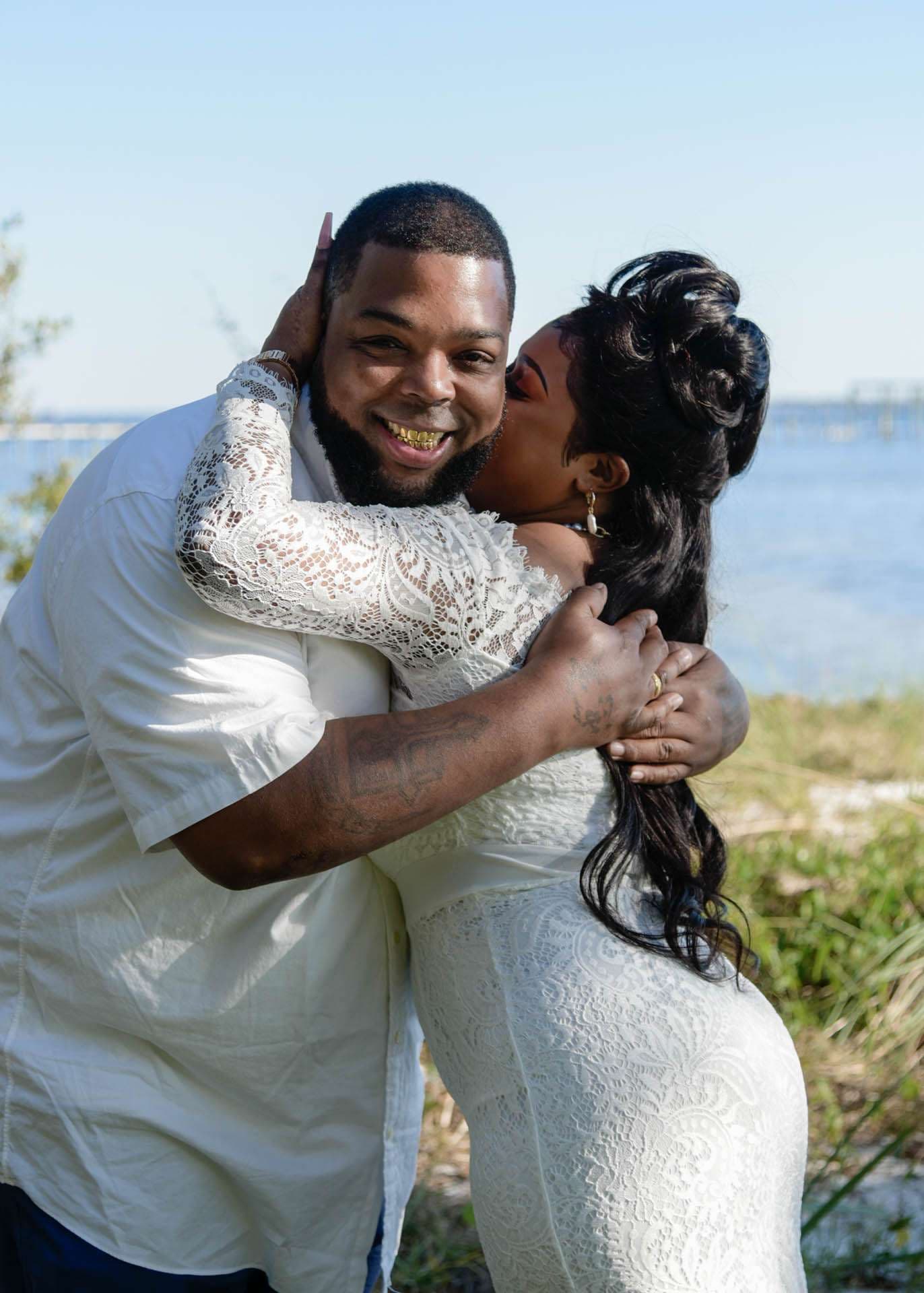 Smiling groom in white shirt embraced by bride in white lace dress kissing his cheek beside the water