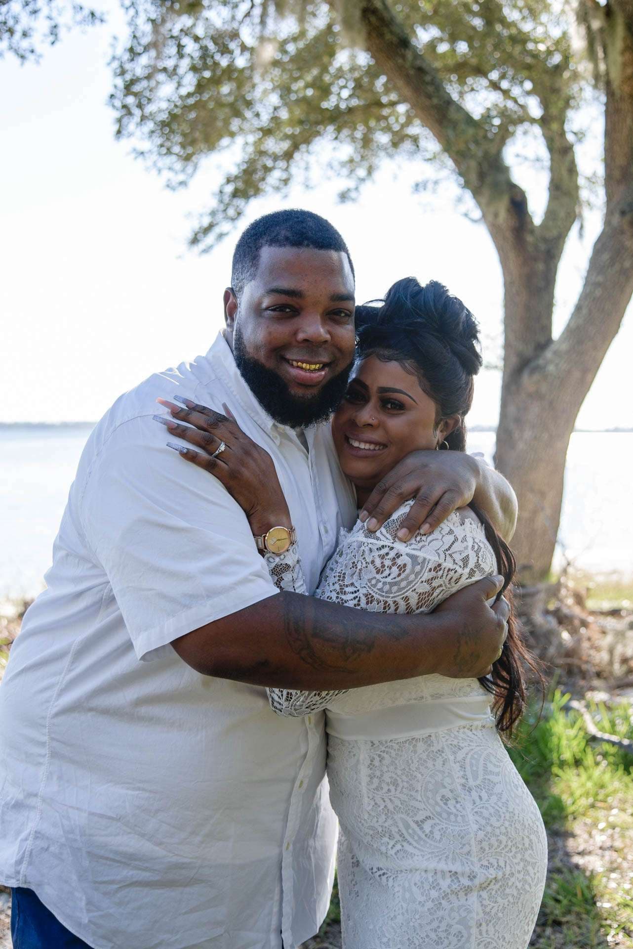 Smiling couple embracing under a live oak tree beside the water, she in a white lace dress, he in a white shirt