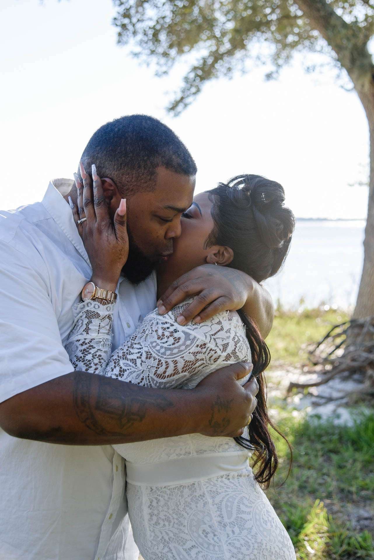 Couple in white embracing and kissing under an oak tree beside the water, her hand on his face