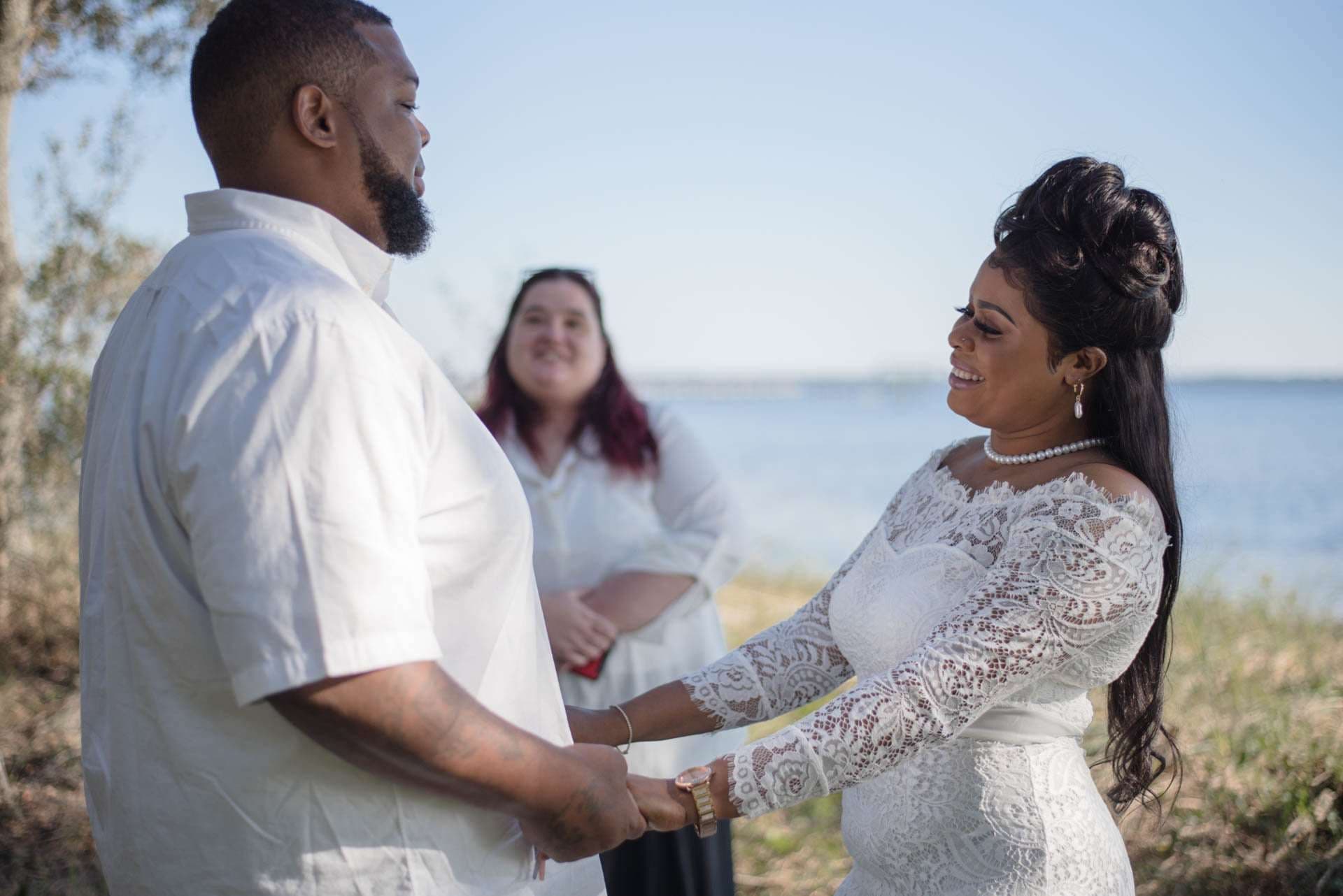 Couple holding hands and smiling during their outdoor ceremony at Oaks By The Bay, officiant smiling behind them