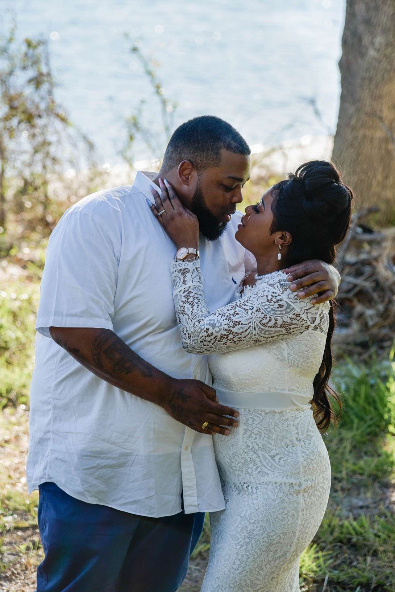 Couple embracing near a tree by the water, she in a white lace dress and he in a white shirt